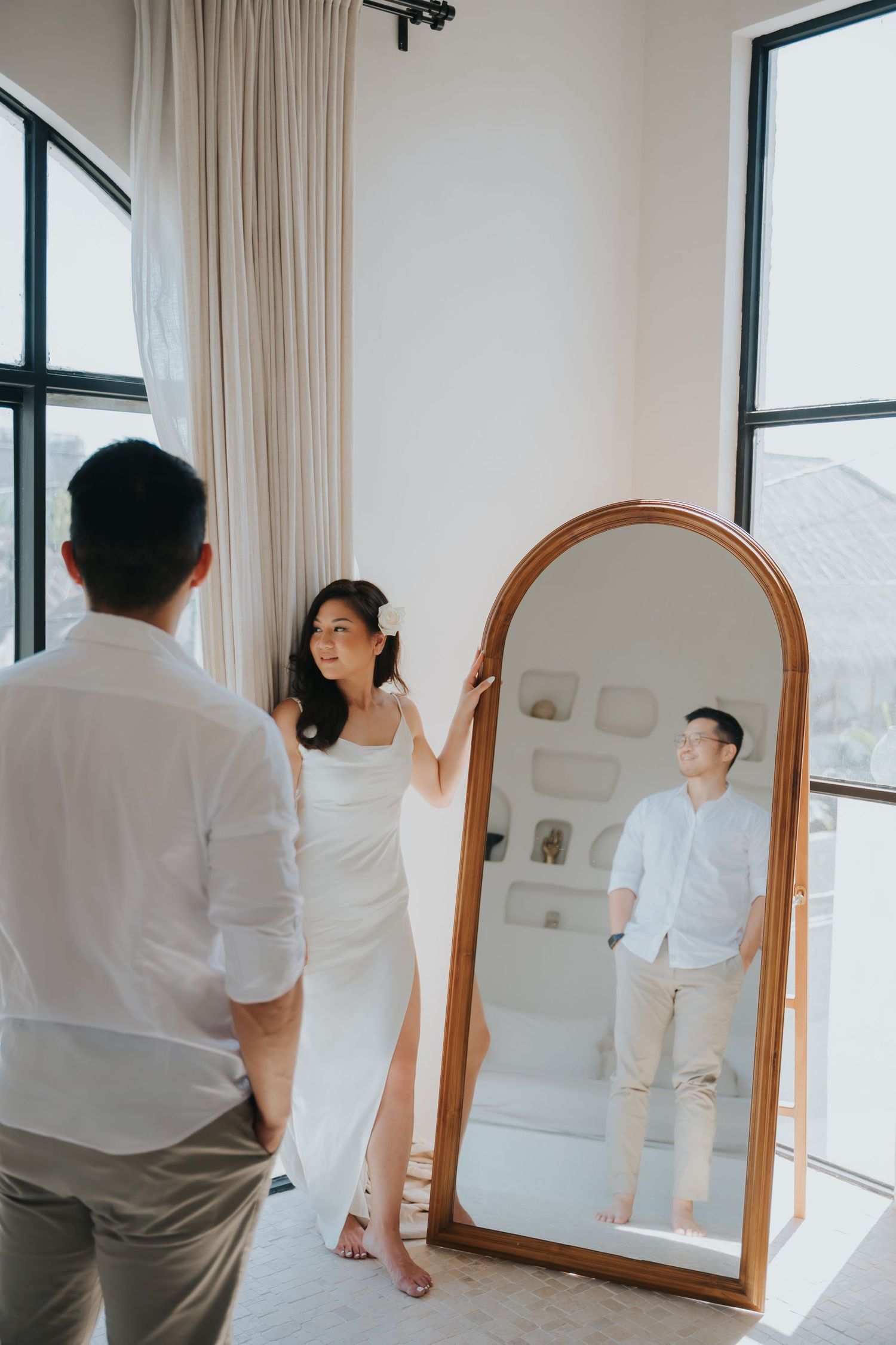 Couple in white outfits posing with an arched wooden floor mirror in a bright minimalist room.