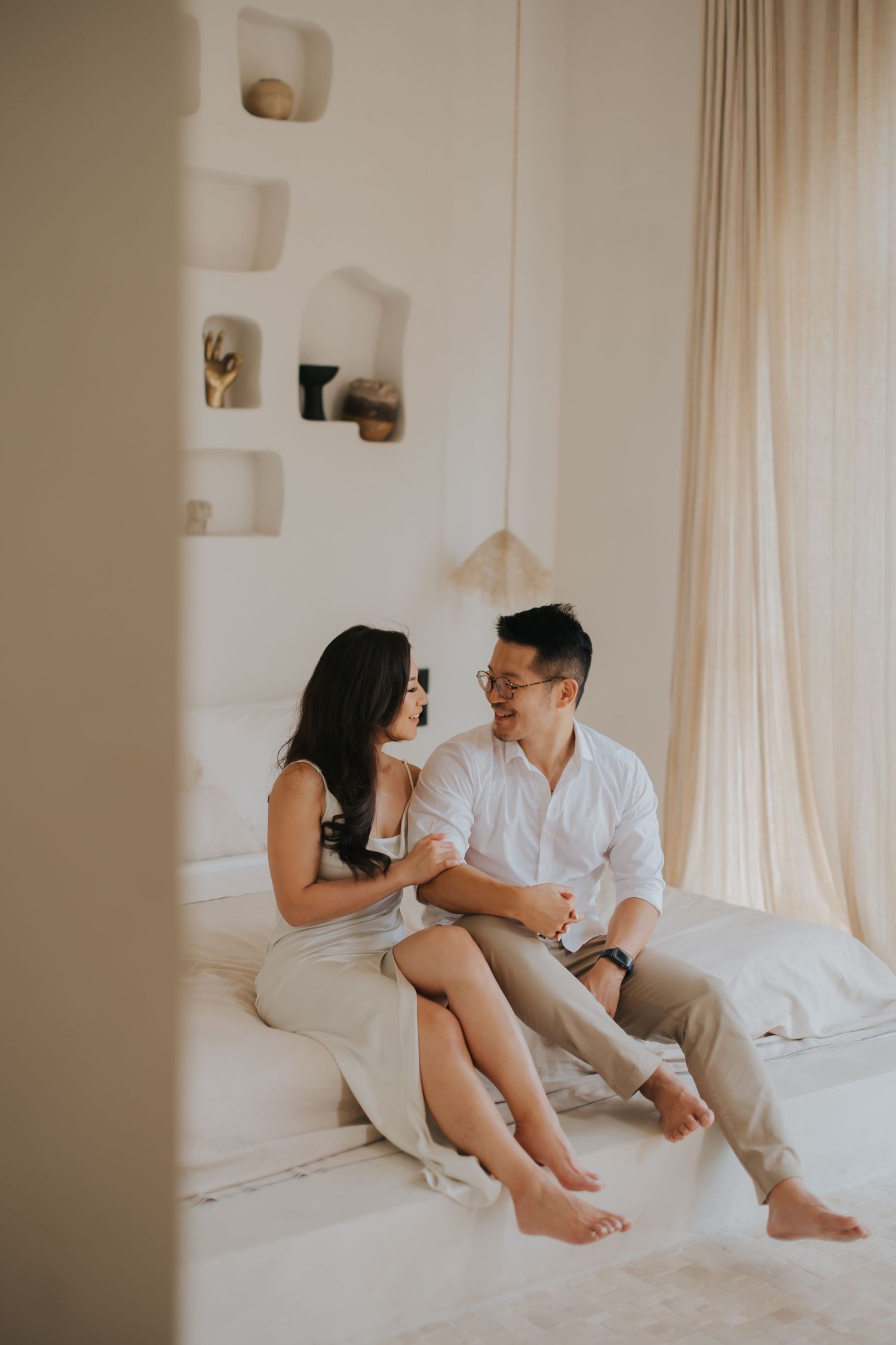 Couple sitting together on a white bed in a bright, minimalist bedroom, smiling and enjoying an intimate conversation.