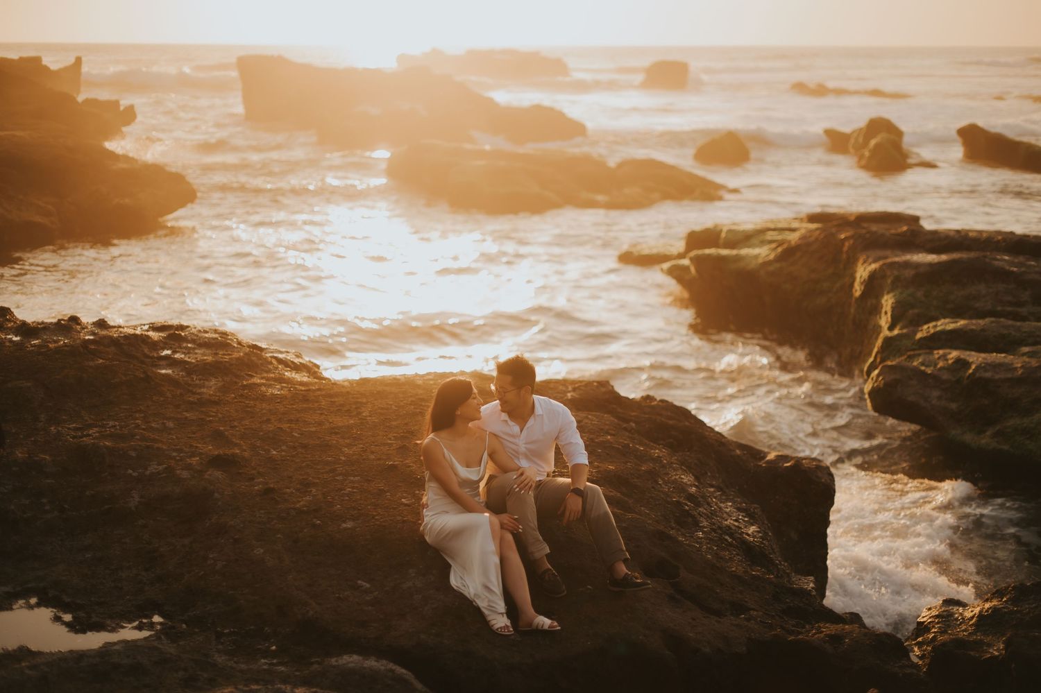 Couple sitting on rocky coastal cliffs at golden sunset, waves crashing behind them in a romantic engagement photo.