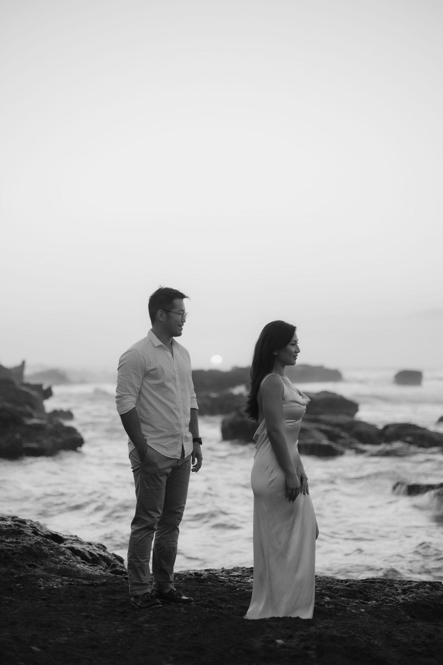 Black and white photo of a couple standing on rocky shore with ocean waves, woman in white dress, man in casual attire.