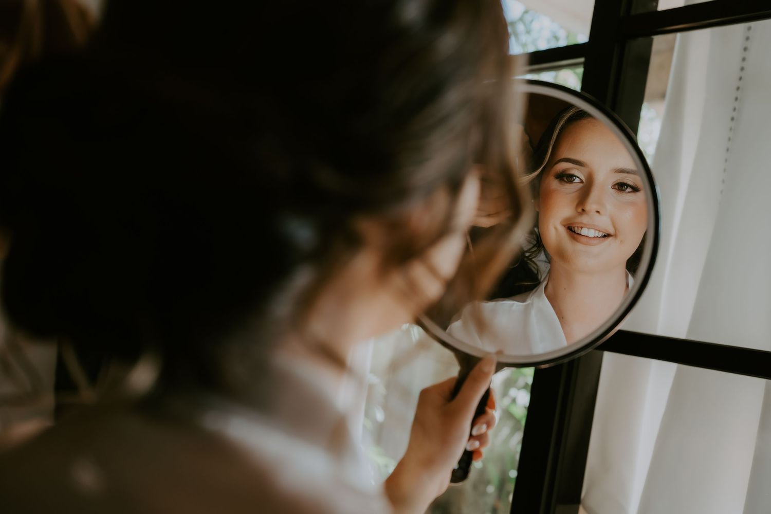 Bride admiring her wedding day look in a round handheld mirror, smiling beautifully before the ceremony.