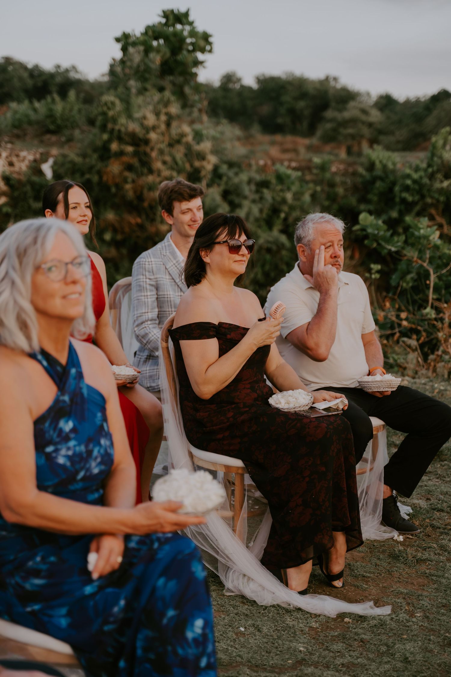 Wedding guests seated outdoors smiling and enjoying a ceremony at sunset in a garden setting.