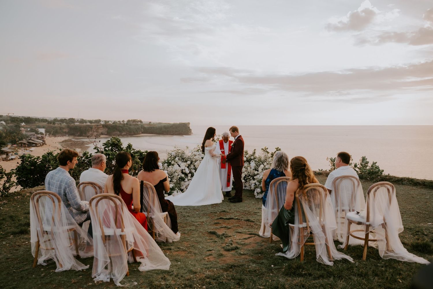 Intimate outdoor wedding ceremony on a cliffside overlooking the ocean at sunset with guests seated in elegant chairs.