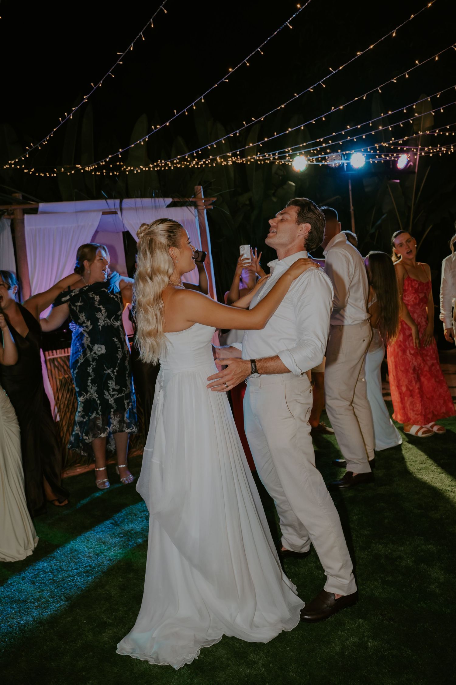 Bride and groom share first dance under twinkling string lights surrounded by guests at an outdoor evening reception.