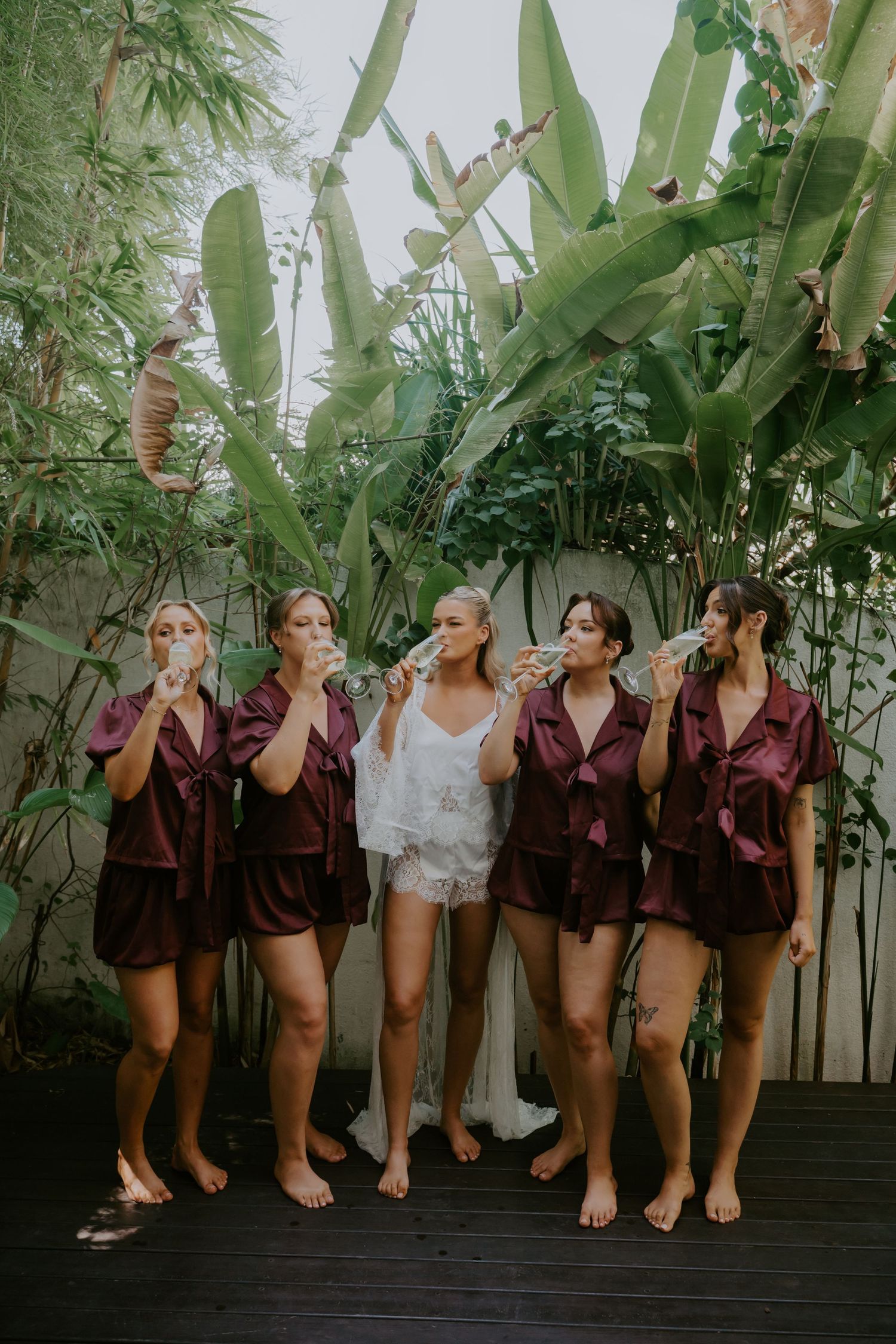 Bridesmaids in matching burgundy satin shorts sets drinking champagne with bride in white lace on tropical deck.