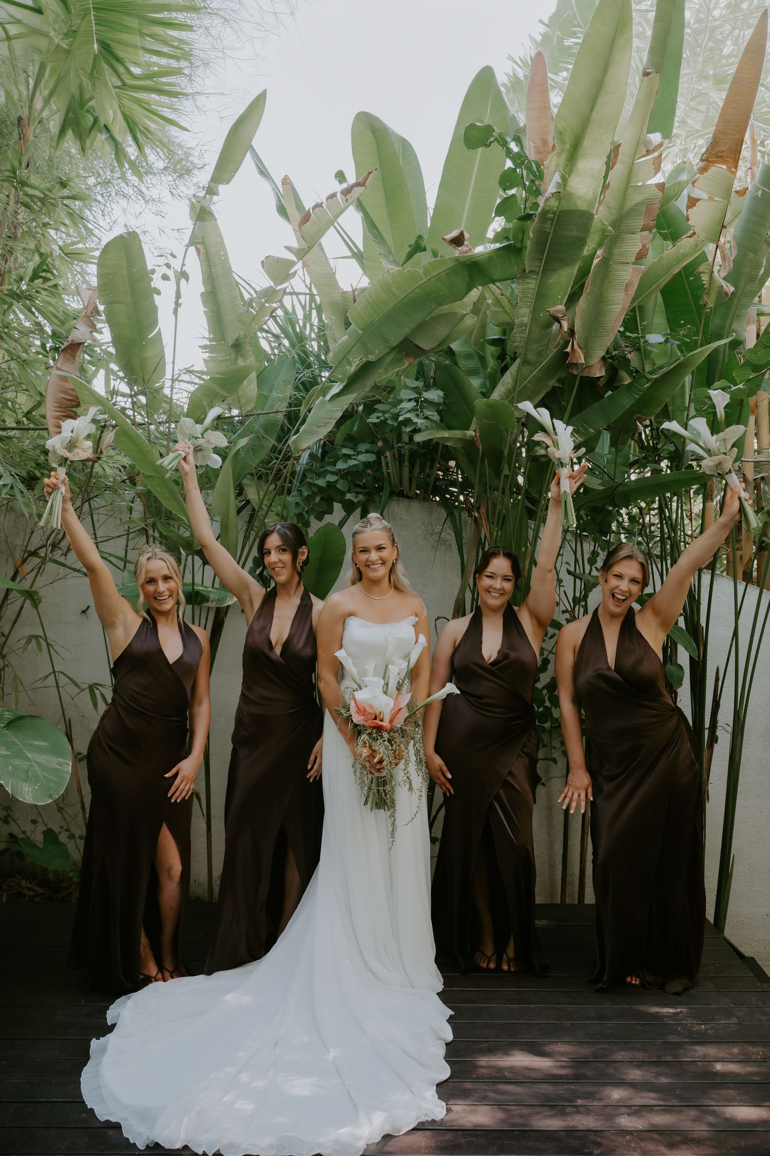 Bride in white gown poses with four bridesmaids in dark brown dresses, raising bouquets before lush tropical greenery.