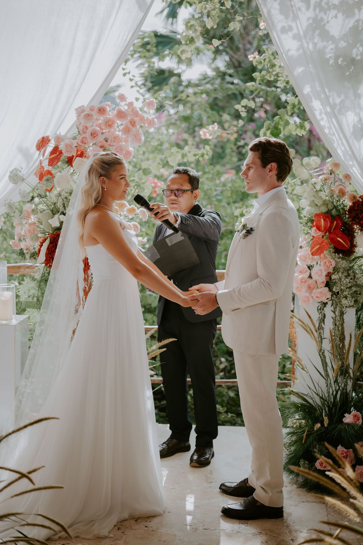 Bride and groom exchange vows holding hands beneath a floral arch with officiant.