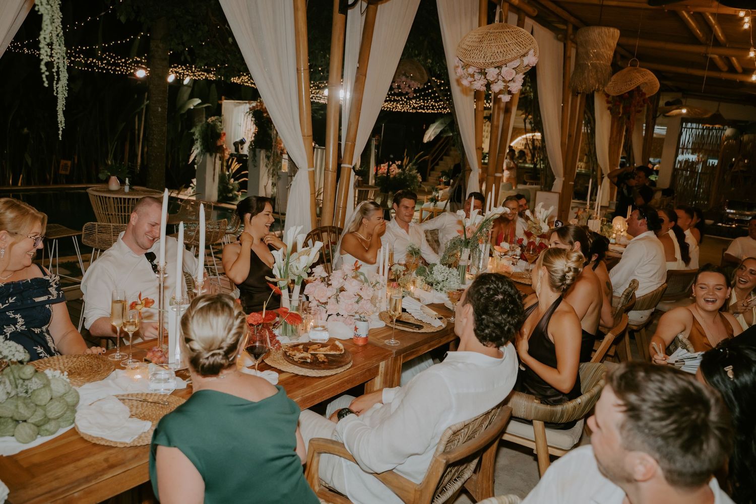 Wedding guests gathered around long decorated dining table during elegant tropical evening reception dinner.