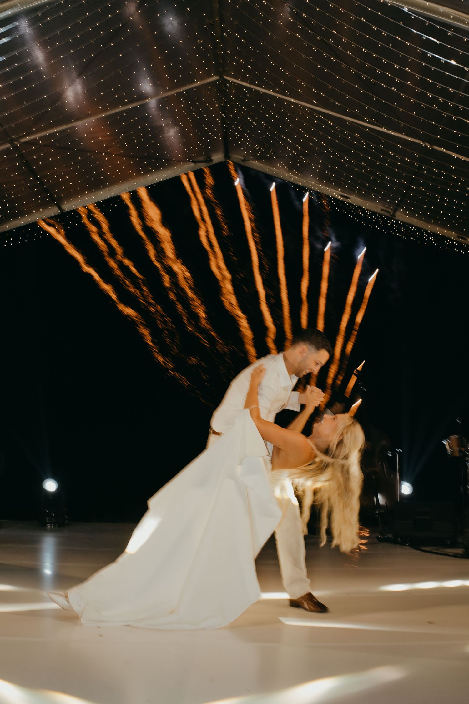 Bride and groom share romantic dip dance under sparkling fairy light canopy with fireworks at wedding.