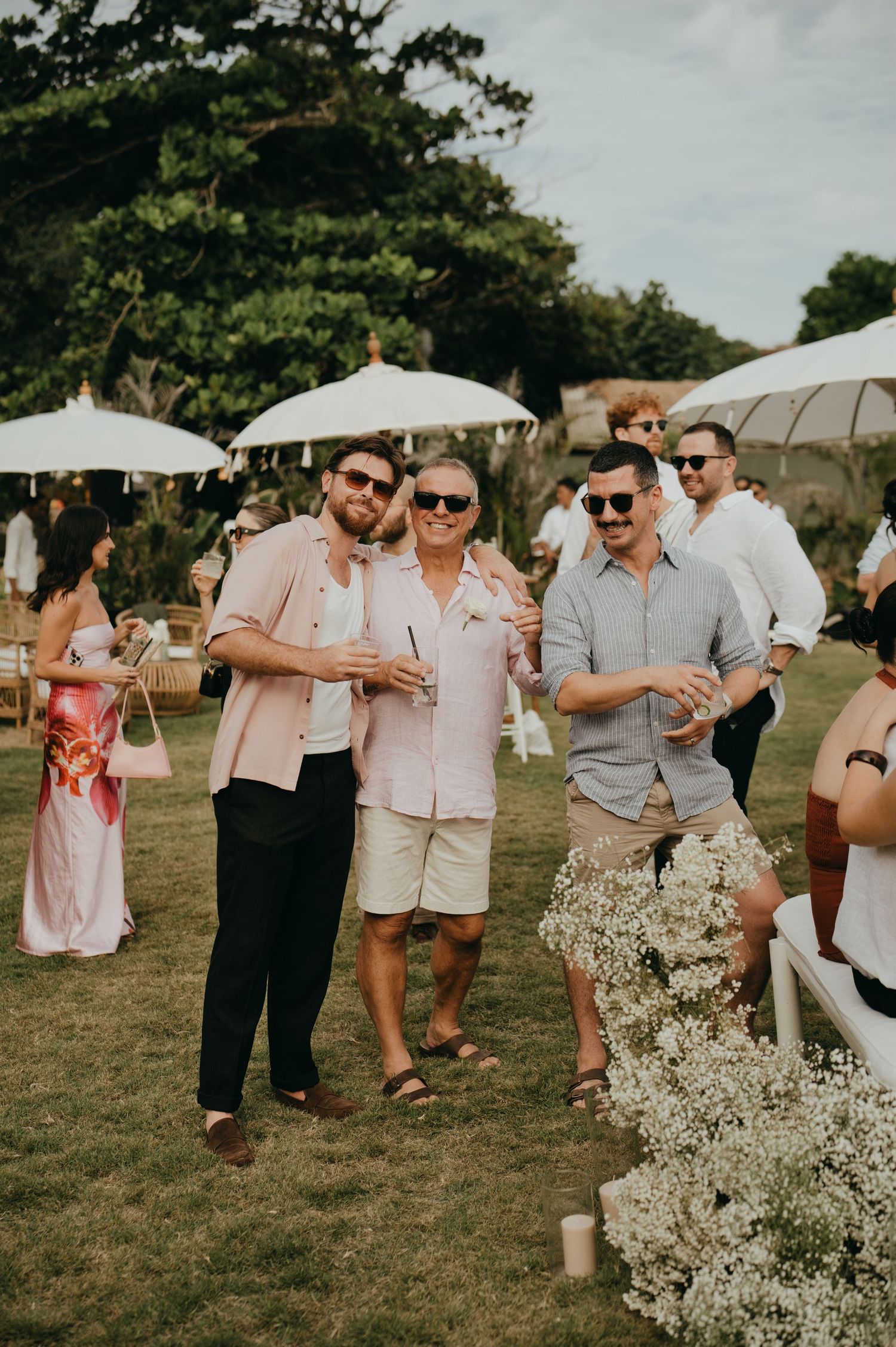 Three men in casual attire smile and hold drinks at an outdoor garden wedding cocktail hour.