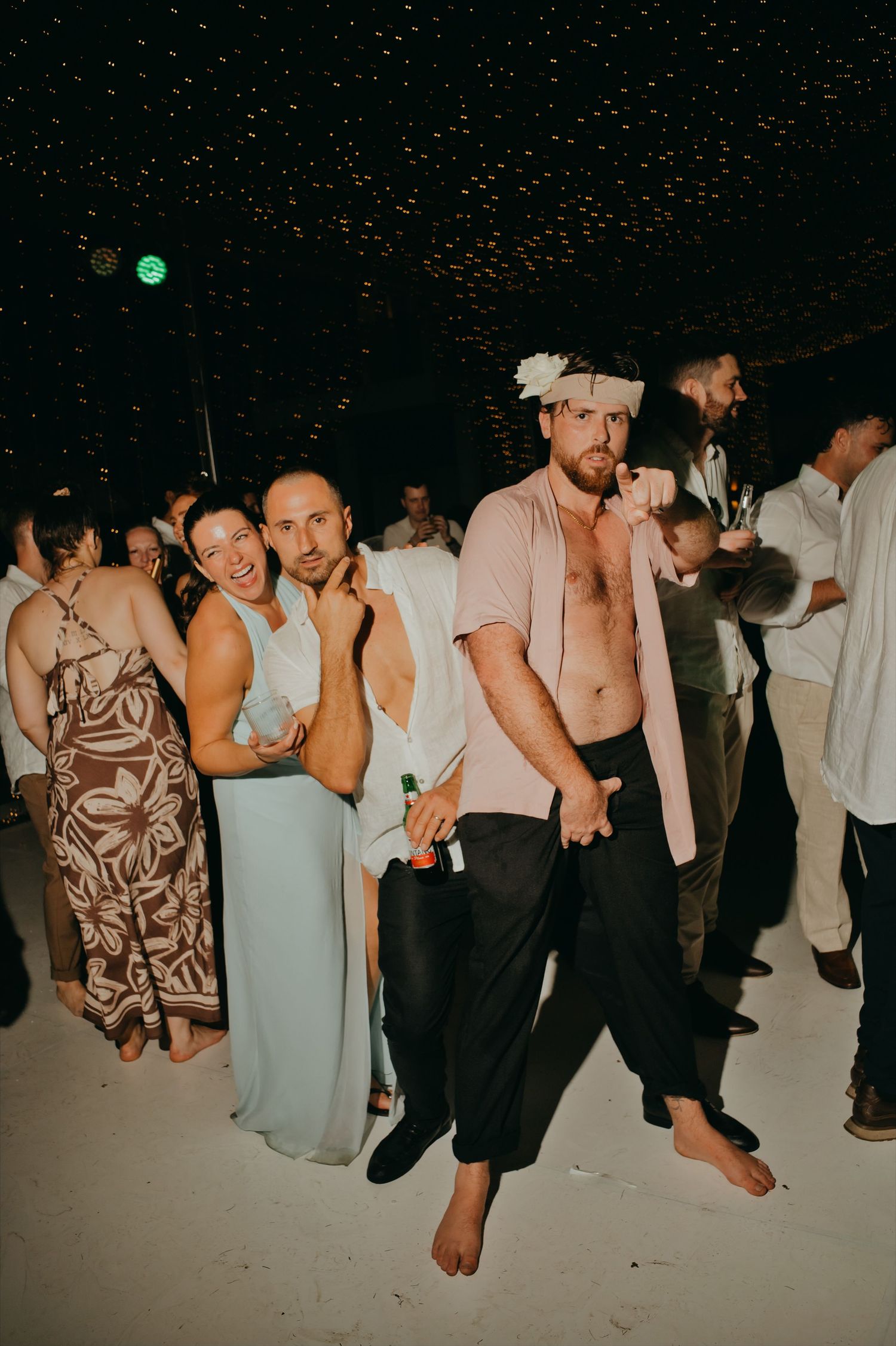 Barefoot wedding guest poses playfully with flower crown at lively nighttime wedding reception dance floor.