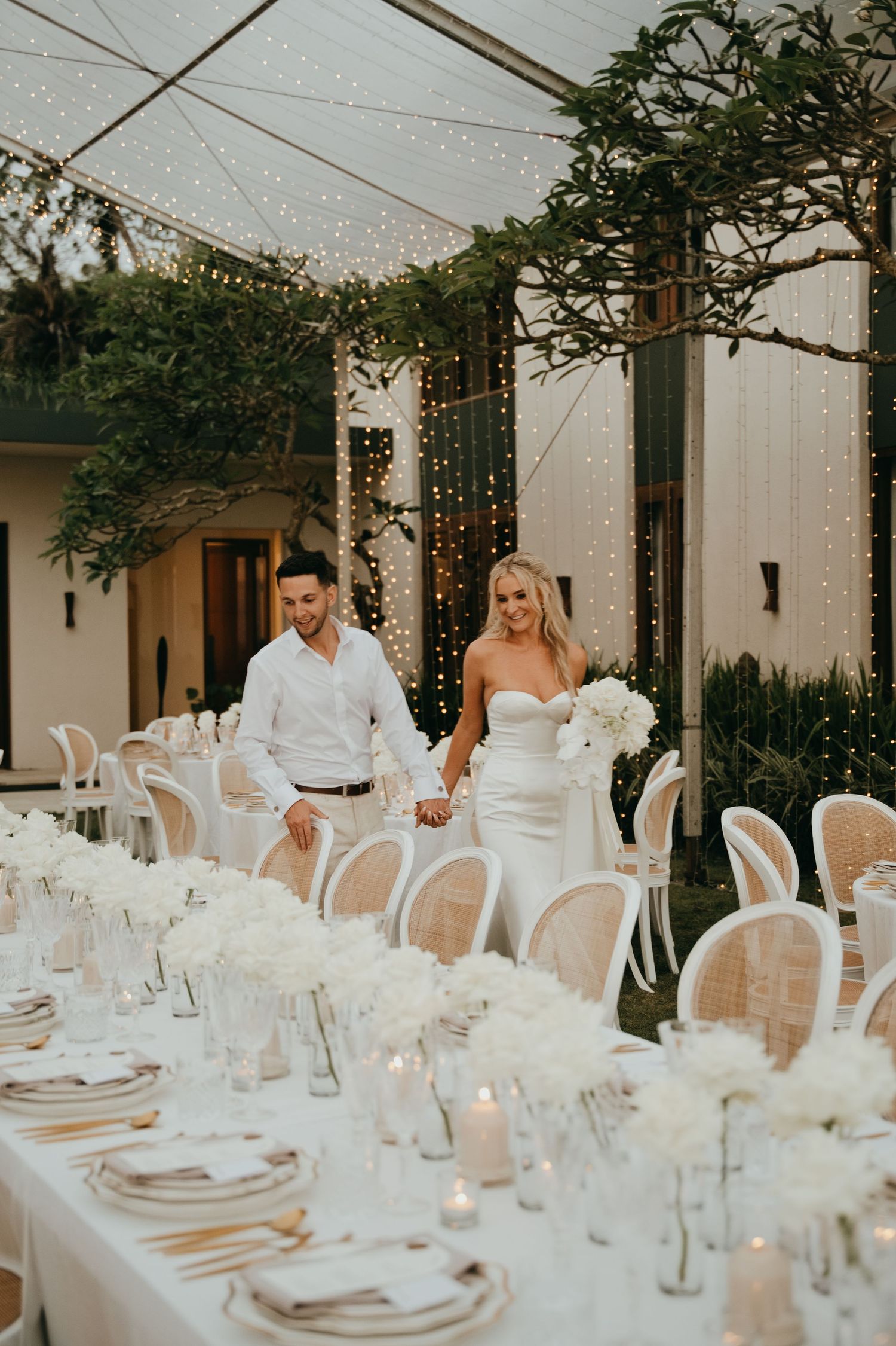 Bride and groom holding hands walking through elegant white reception dinner setup with fairy lights.
