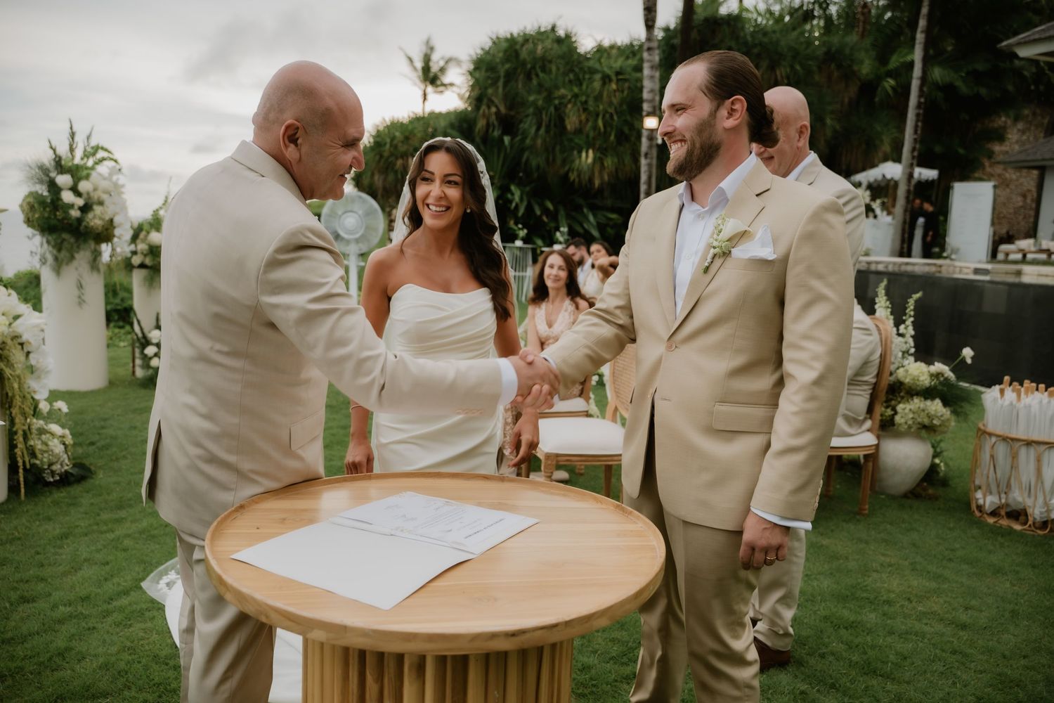 Bride and groom shake hands with officiant at outdoor wedding ceremony beside a wooden signing table.