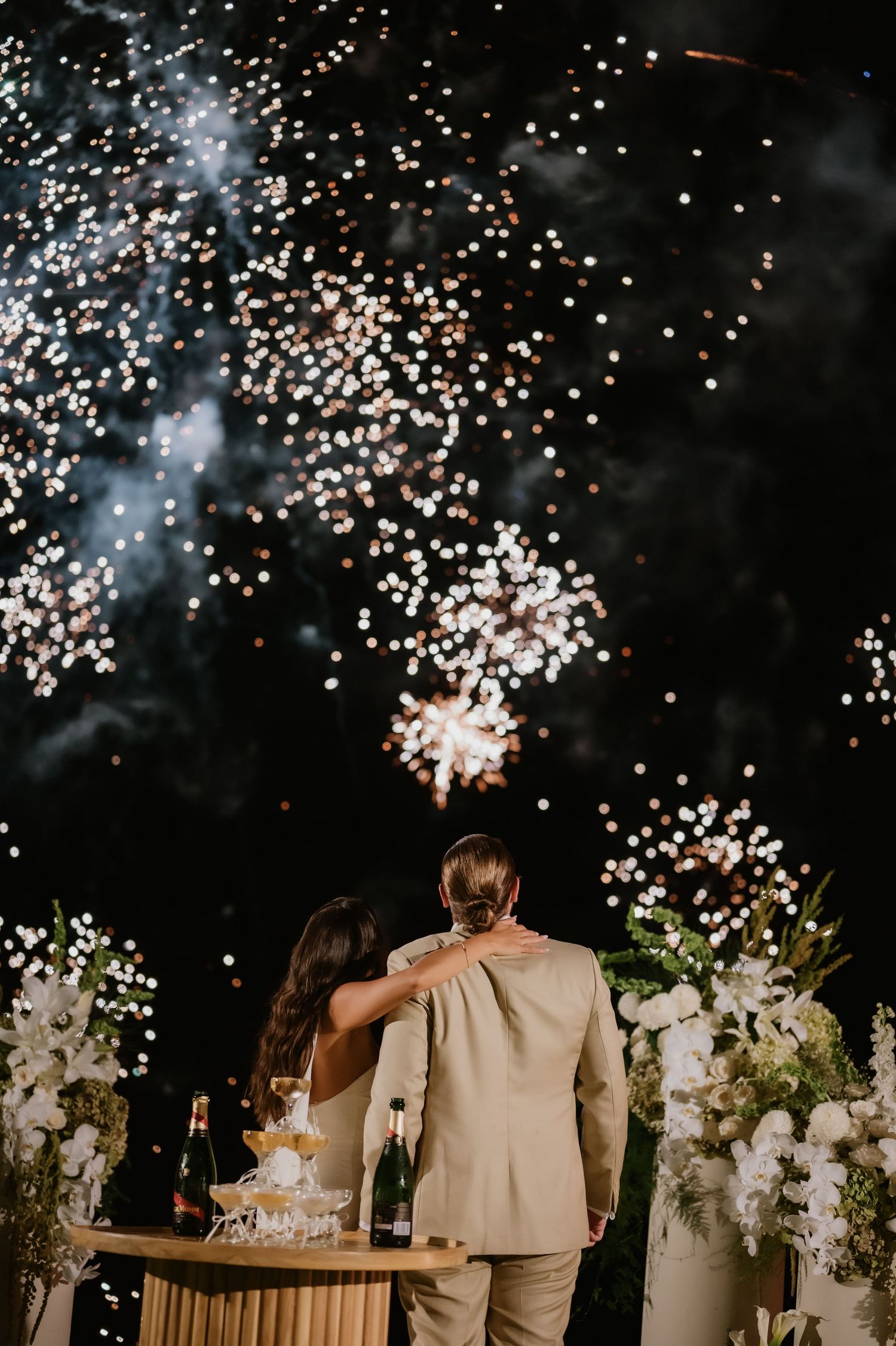 Bride and groom watch stunning fireworks display together at their outdoor wedding reception with white floral arrangements.