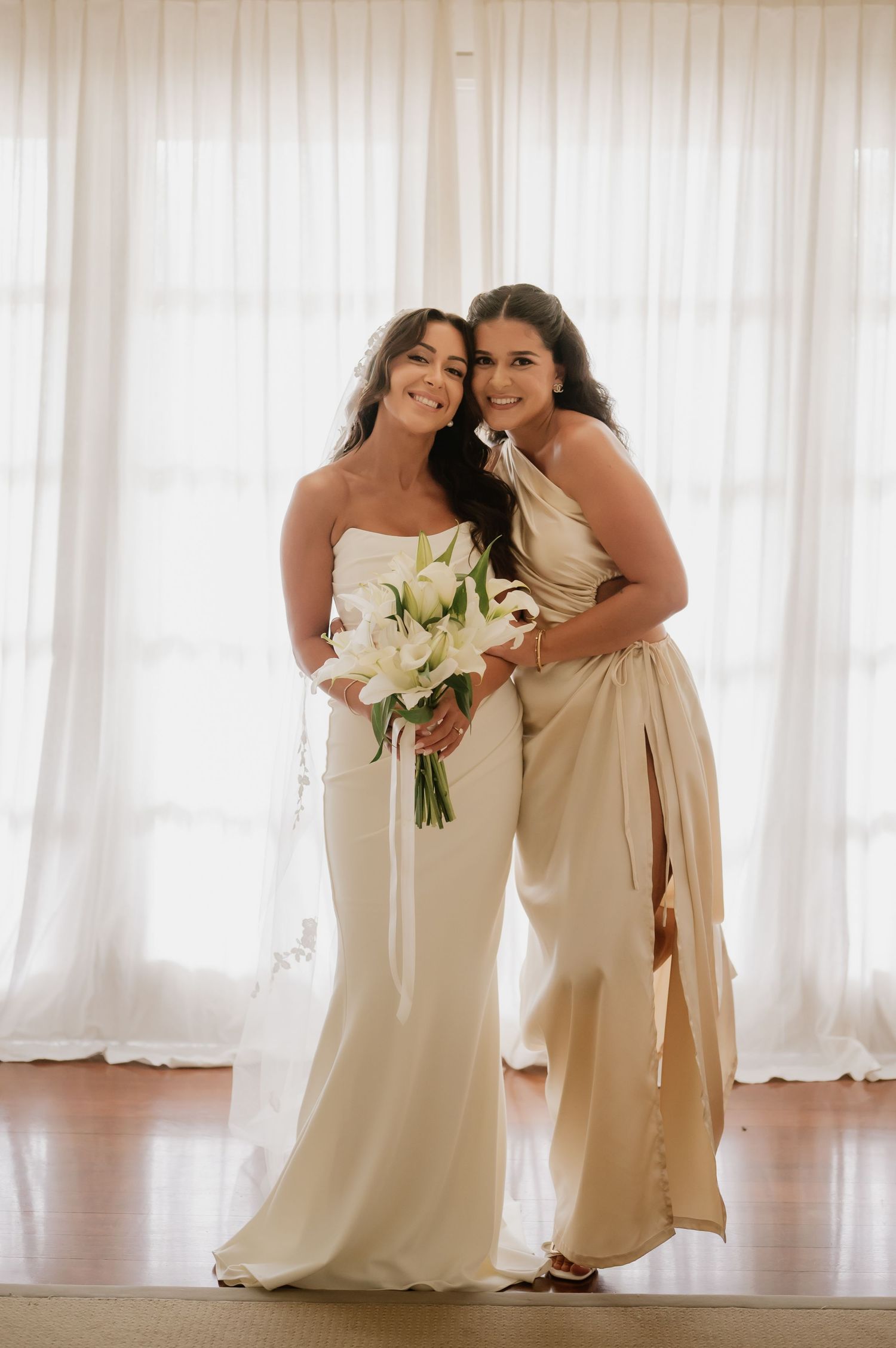 Bride in strapless white gown holding white lily bouquet posing with bridesmaid in champagne dress.