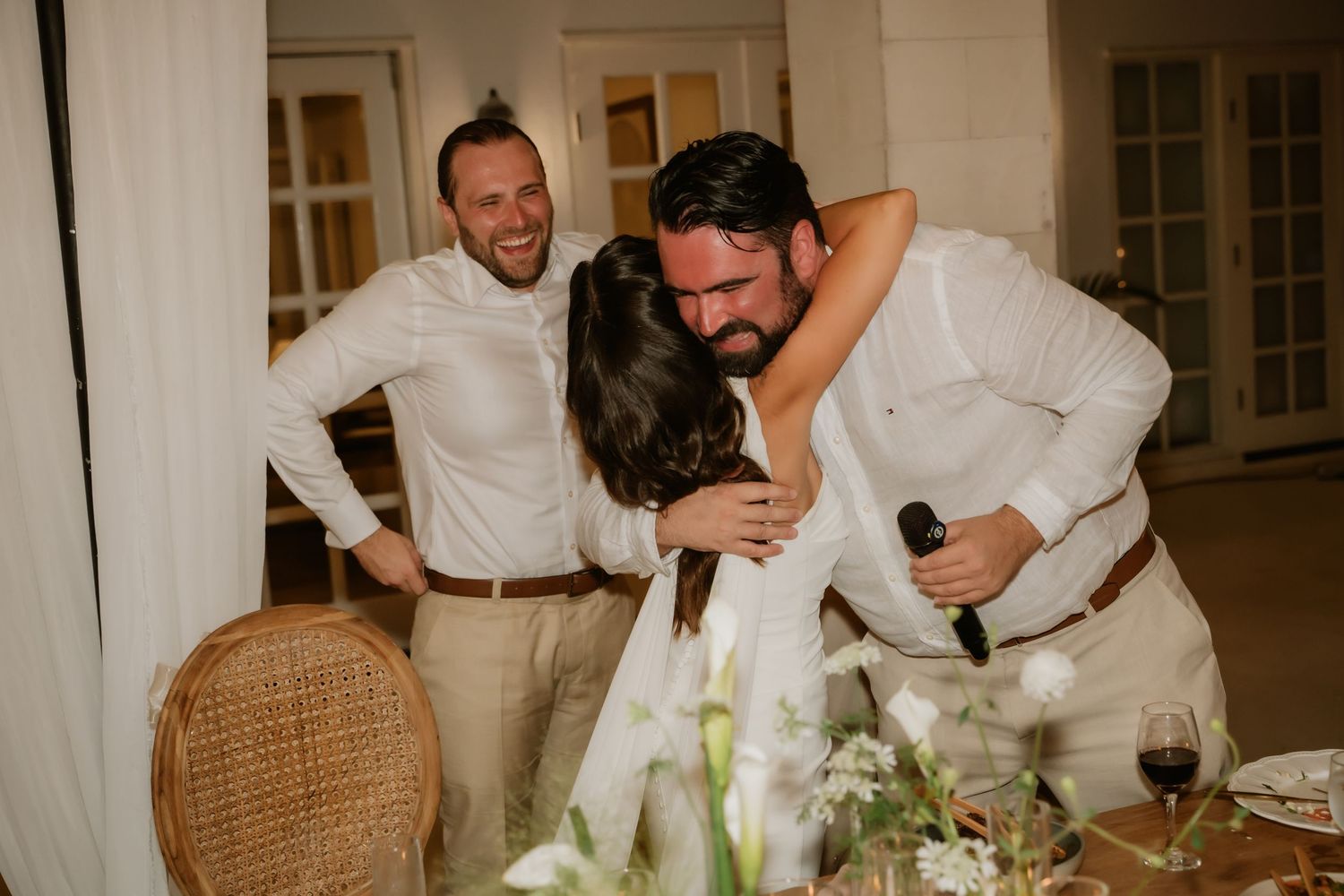 Emotional wedding reception moment as groom embraces bride while groomsman laughs in background.