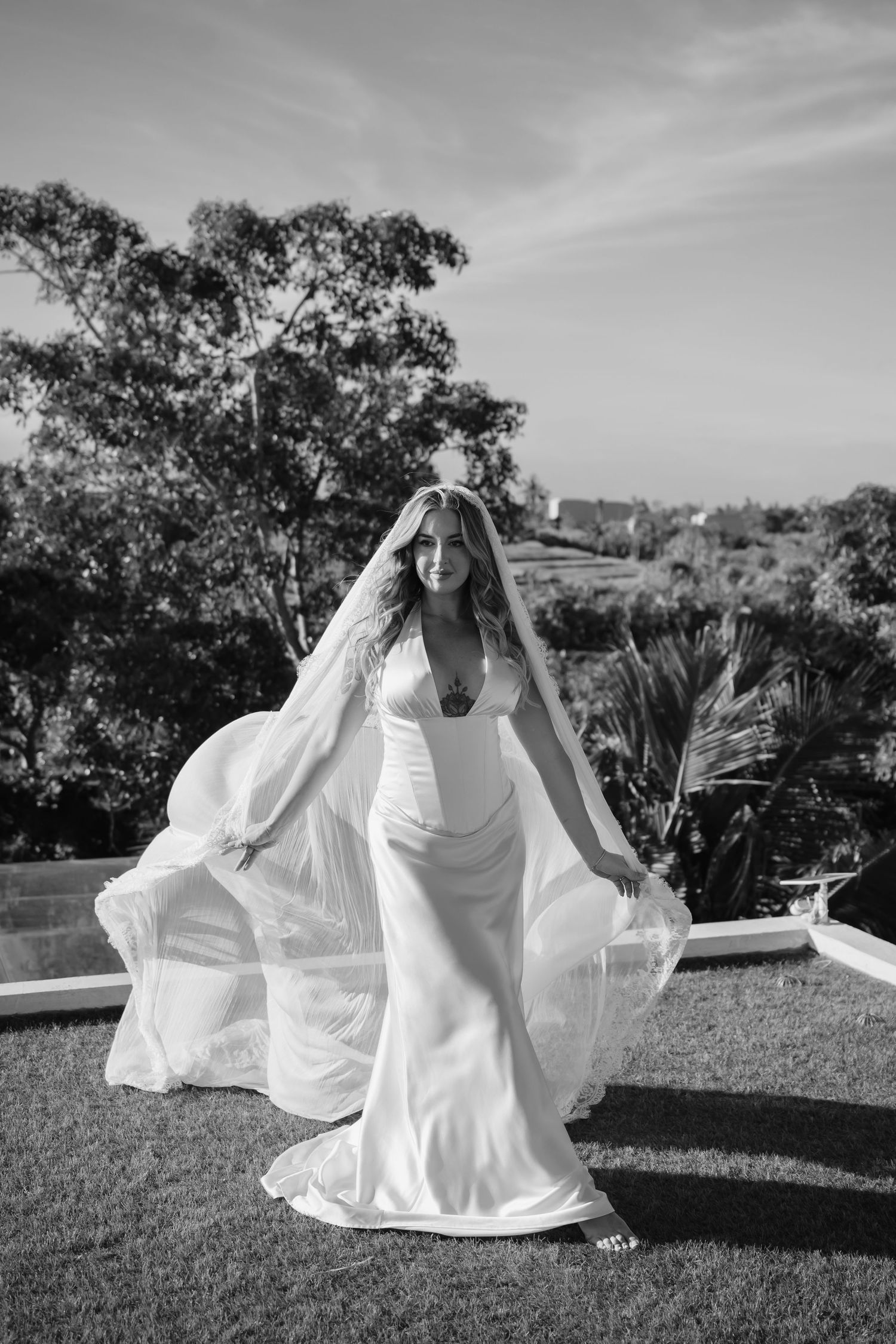 Bride in elegant white gown and flowing veil poses outdoors with tropical trees in black and white wedding photo.
