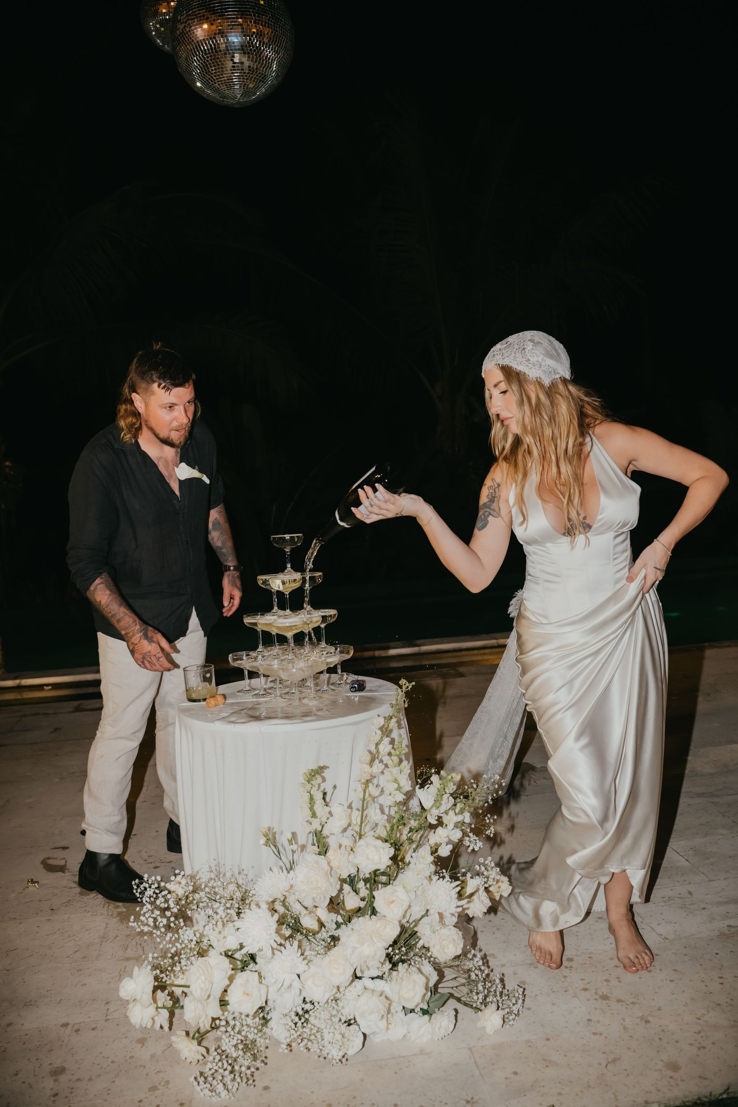 Bride and groom pour champagne tower surrounded by white floral arrangements at outdoor wedding.