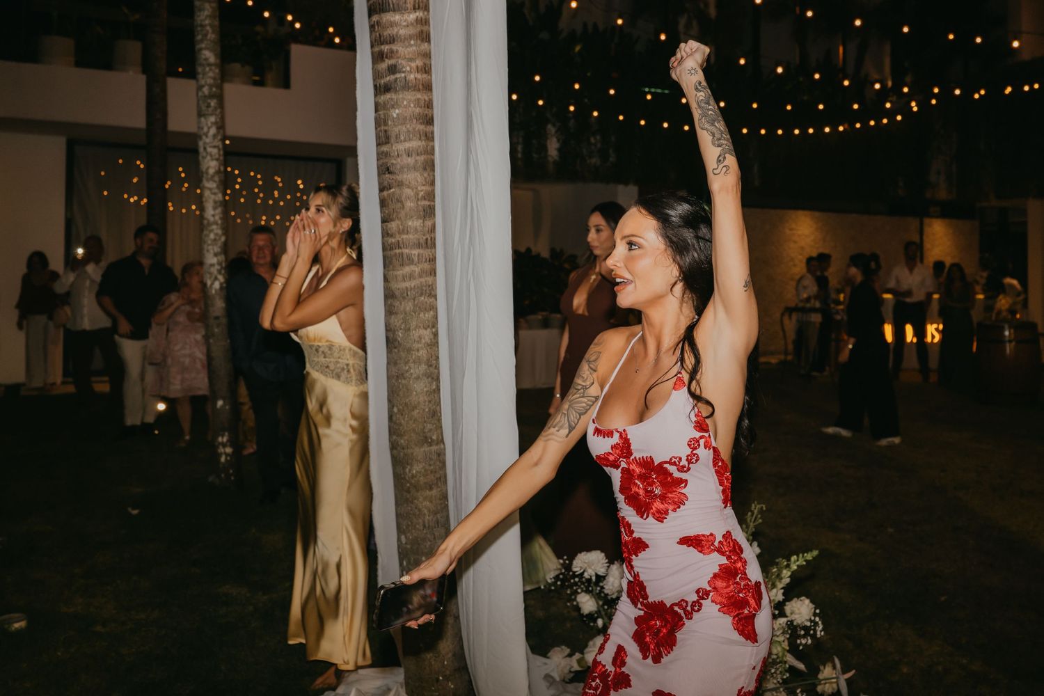 Woman in red floral dress dancing energetically near a palm tree at an outdoor evening wedding reception.