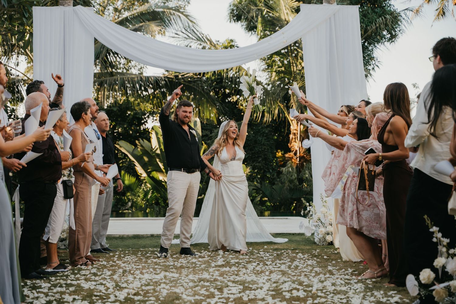 Bride and groom walk down petal-lined aisle under white floral arch as guests celebrate with confetti.