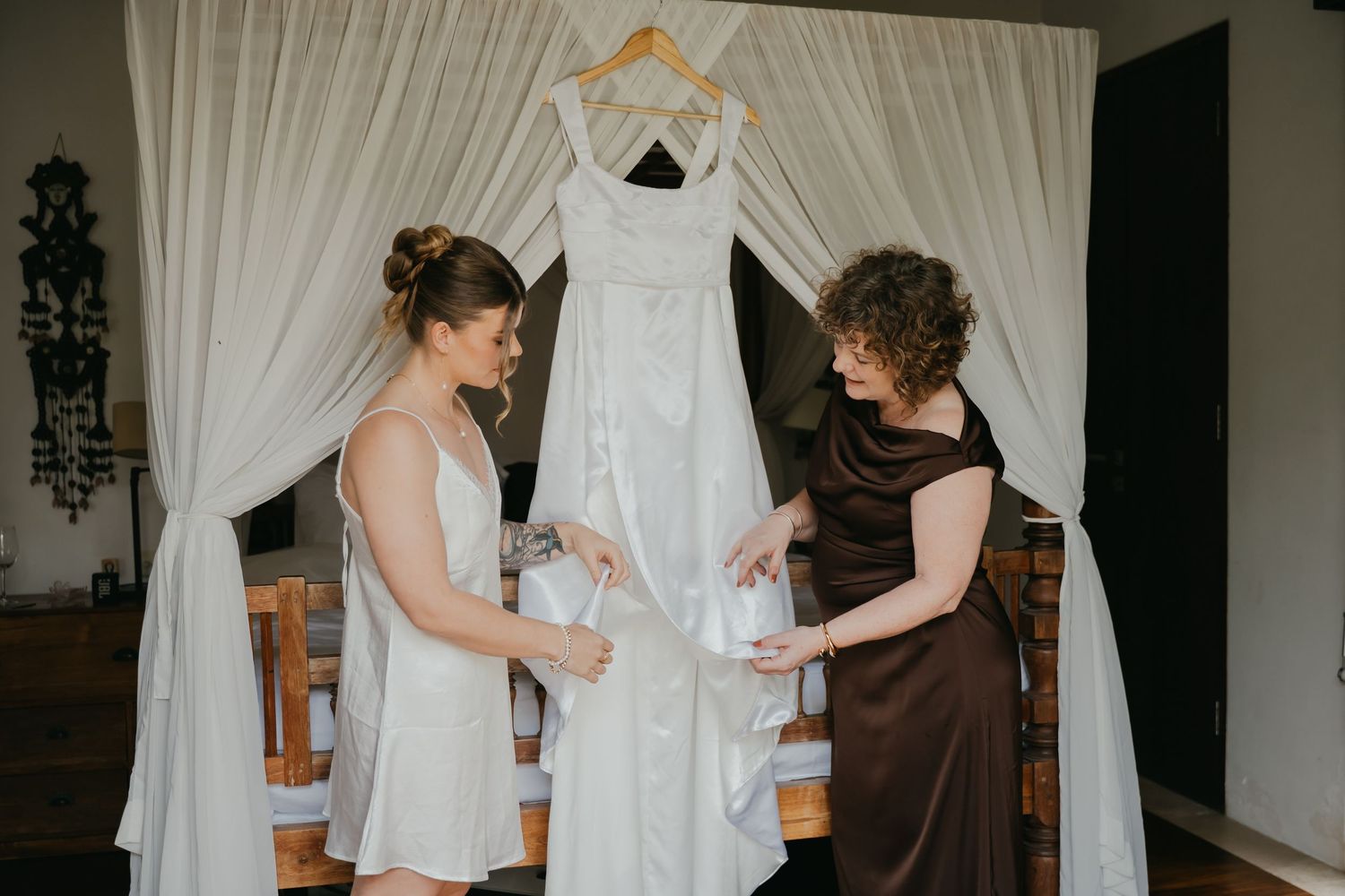 Bride and helper examining white wedding dress hanging on canopy bed frame before ceremony.