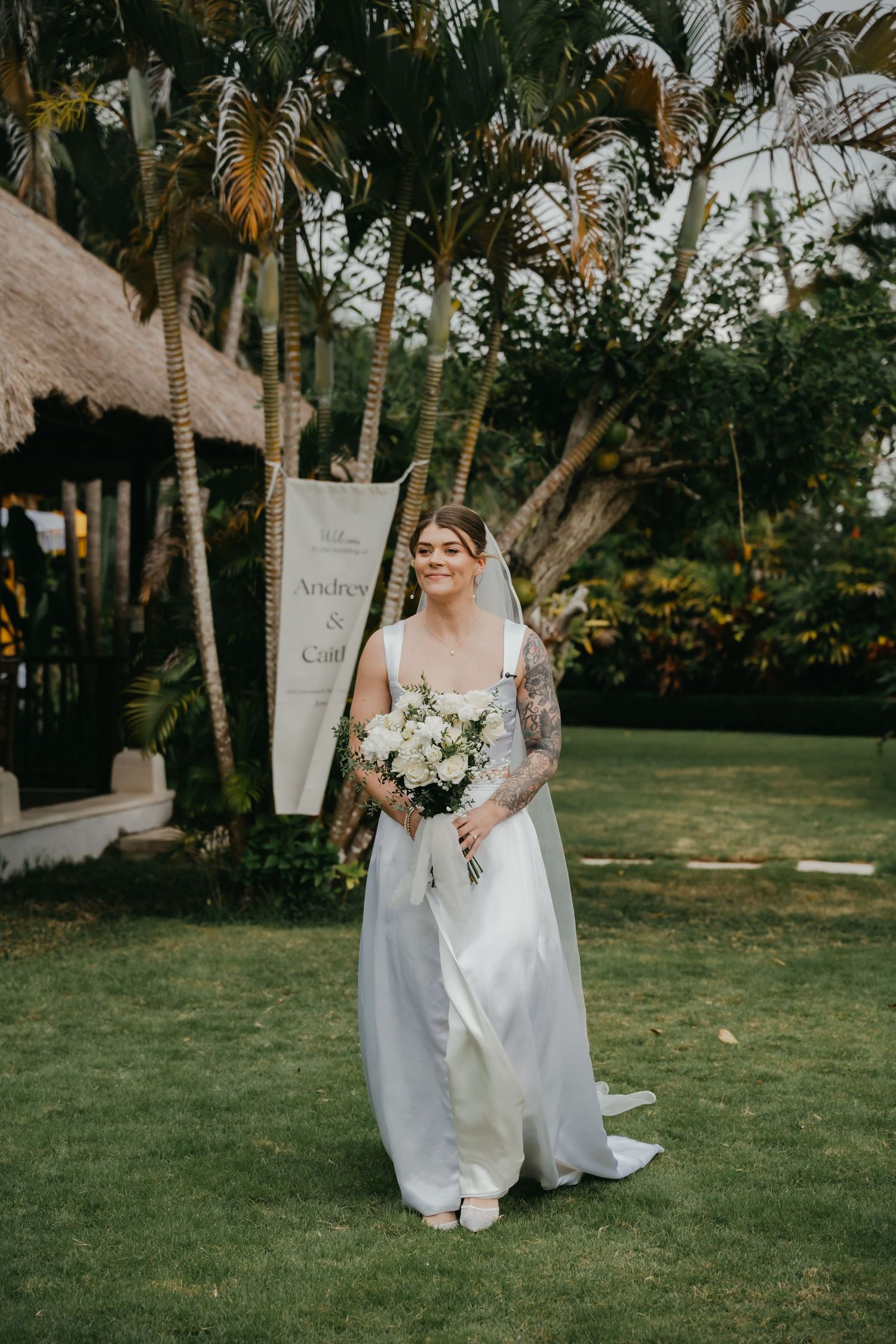 Smiling bride with tattoos wearing white two-piece wedding gown holds white bouquet in tropical garden setting.