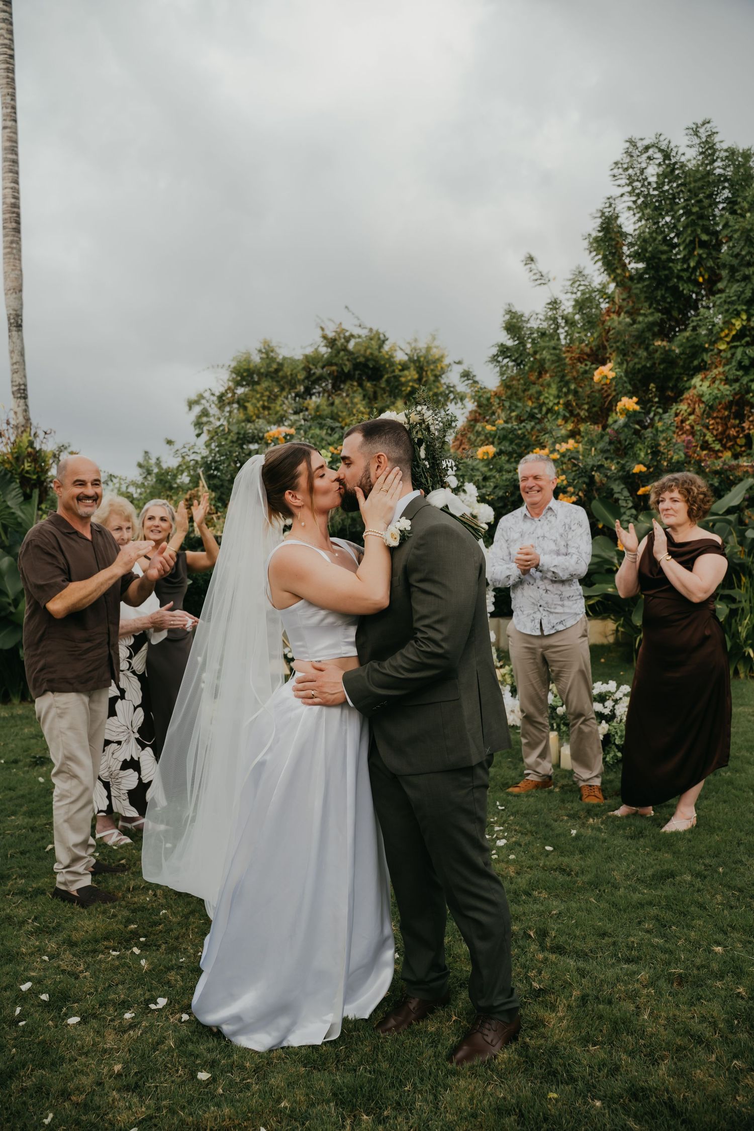 Bride and groom share first kiss as married couple while guests cheer and applaud outdoors.
