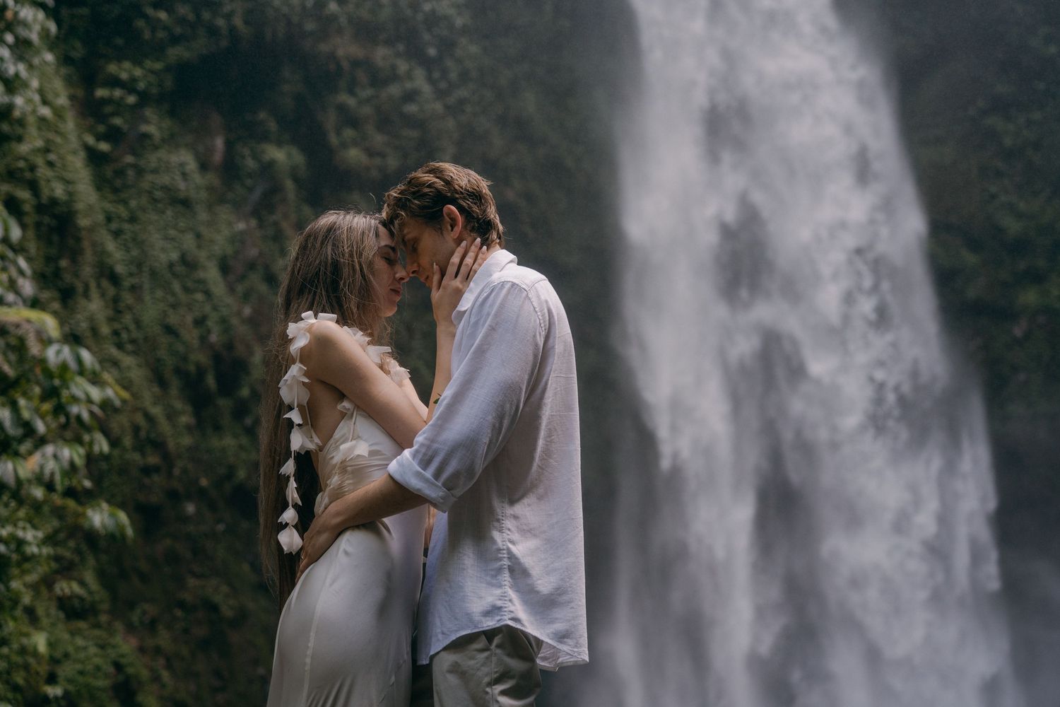 A romantic couple embraces in front of a majestic waterfall in a lush forest setting.