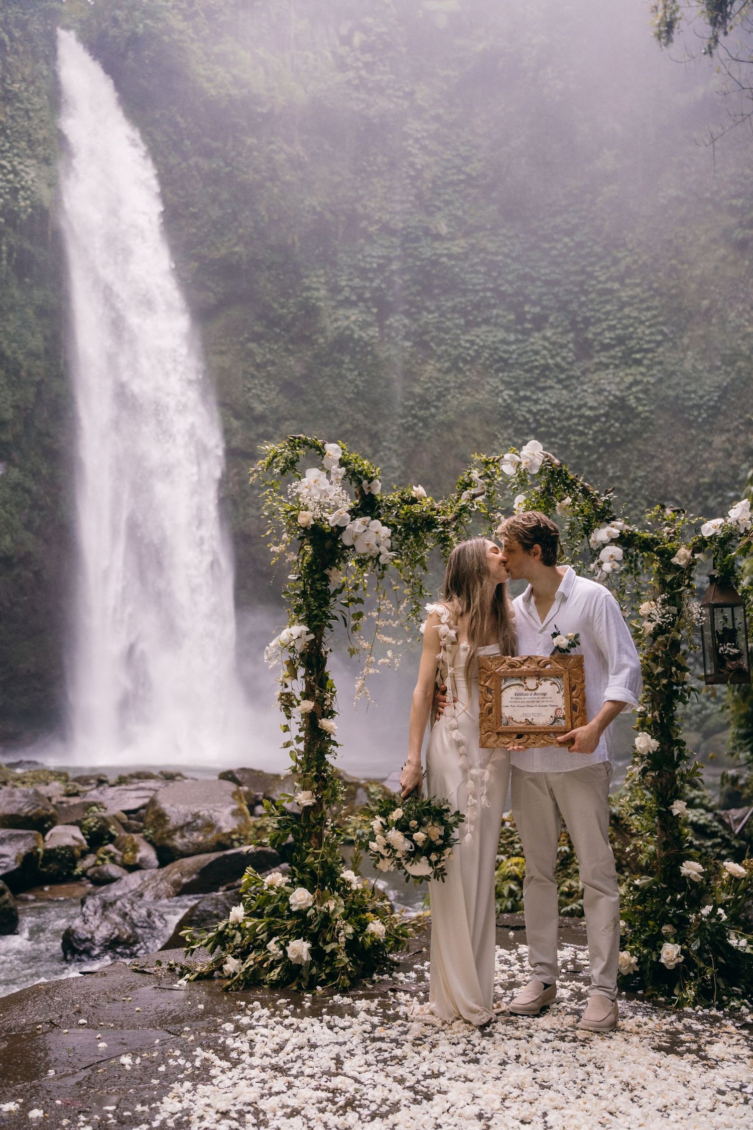 A couple shares an intimate moment in front of a majestic waterfall surrounded by a floral arch and misty atmosphere.