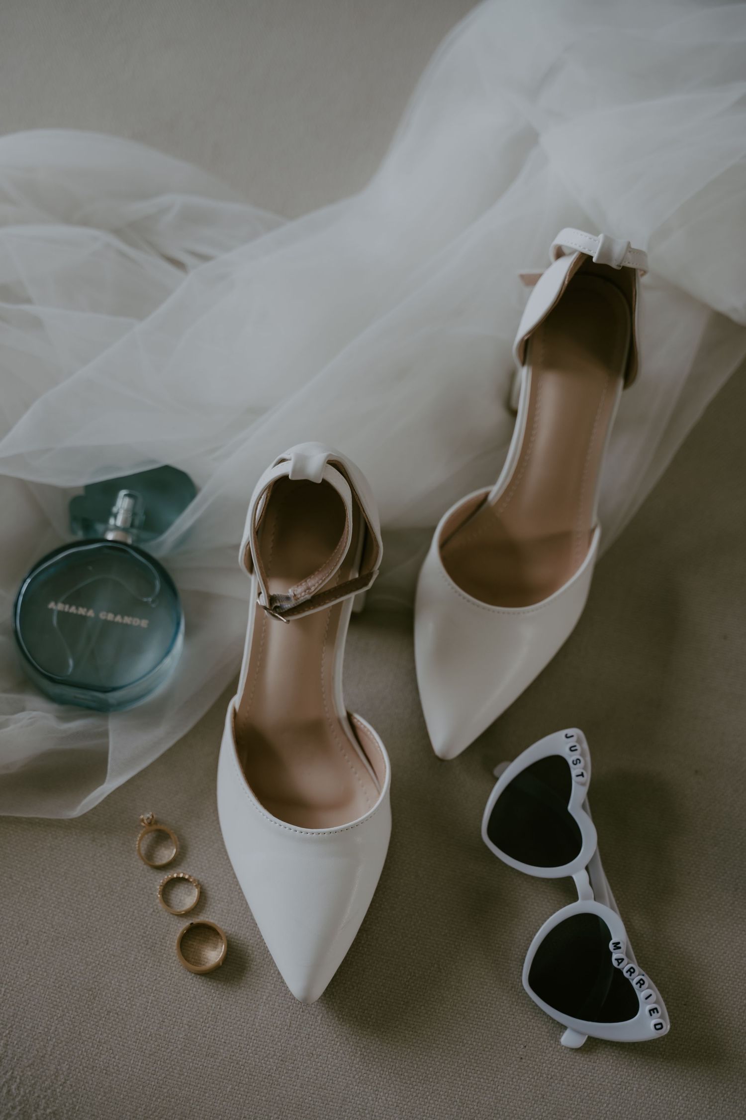 Wedding accessories including white heels, sunglasses, and rings arranged on neutral background.