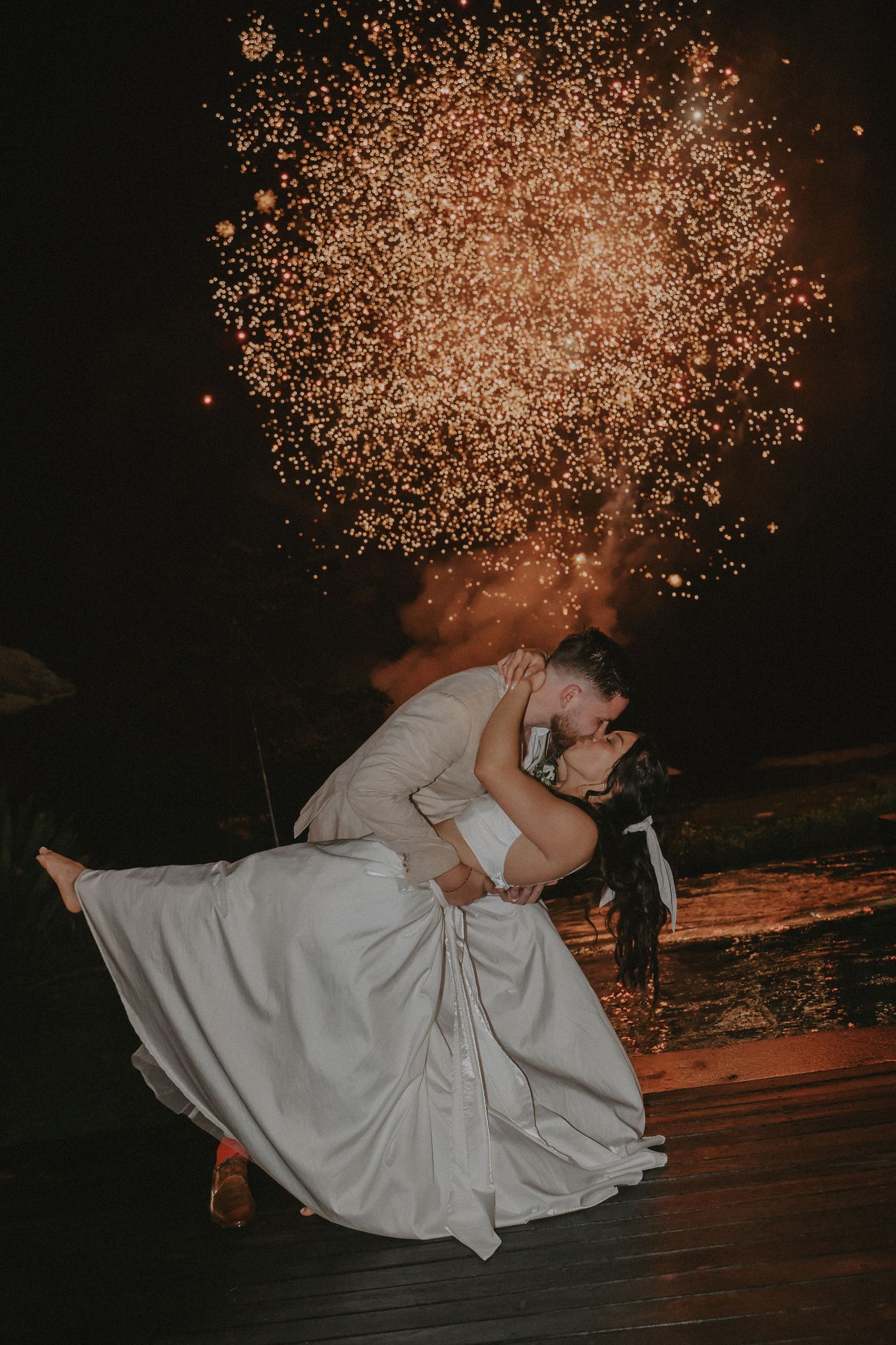 A romantic couple in wedding attire dips and kisses under a shower of golden sparklers at night.