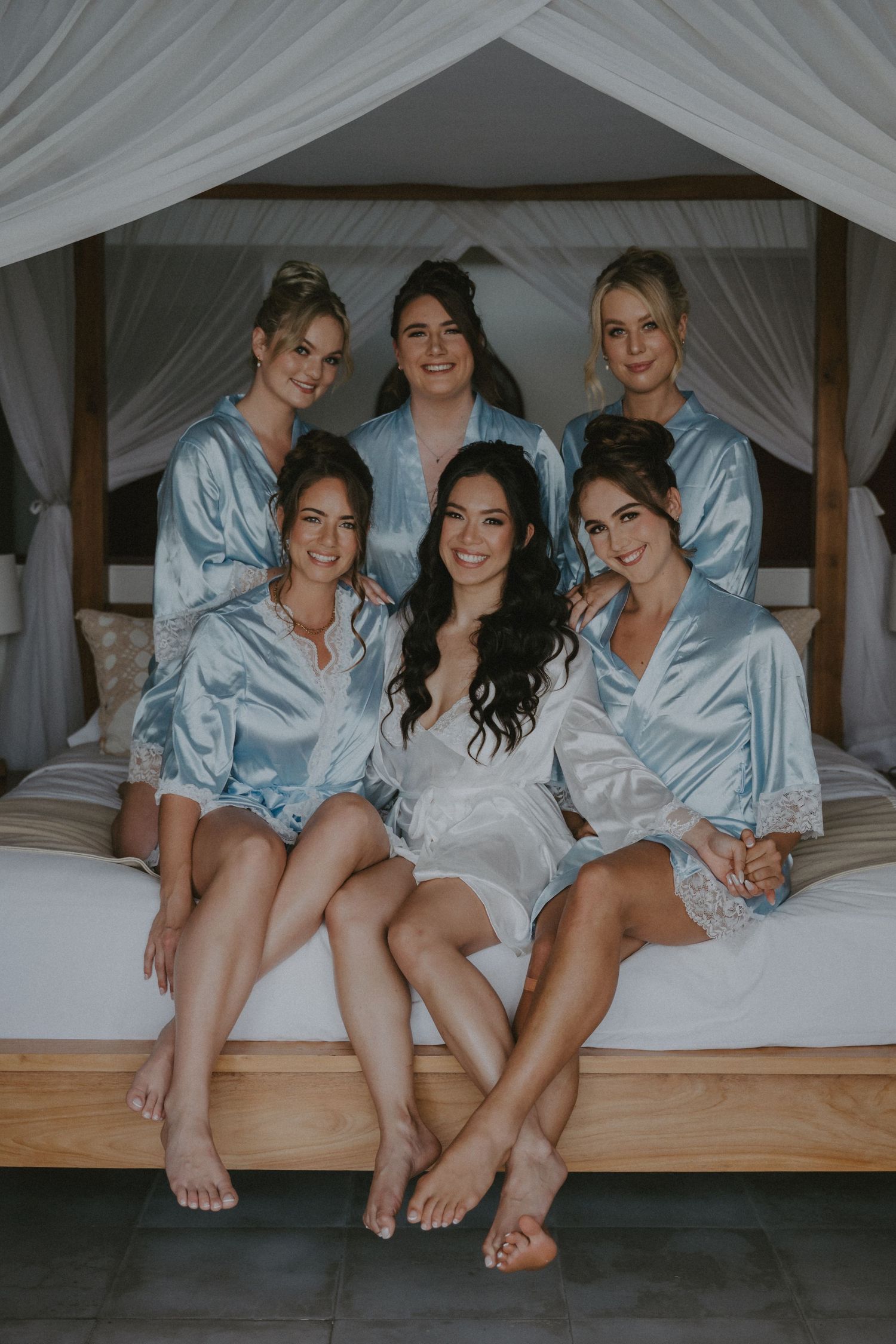 A group in matching light blue satin robes and one in white sit together on a canopy bed for a pre-wedding photo.