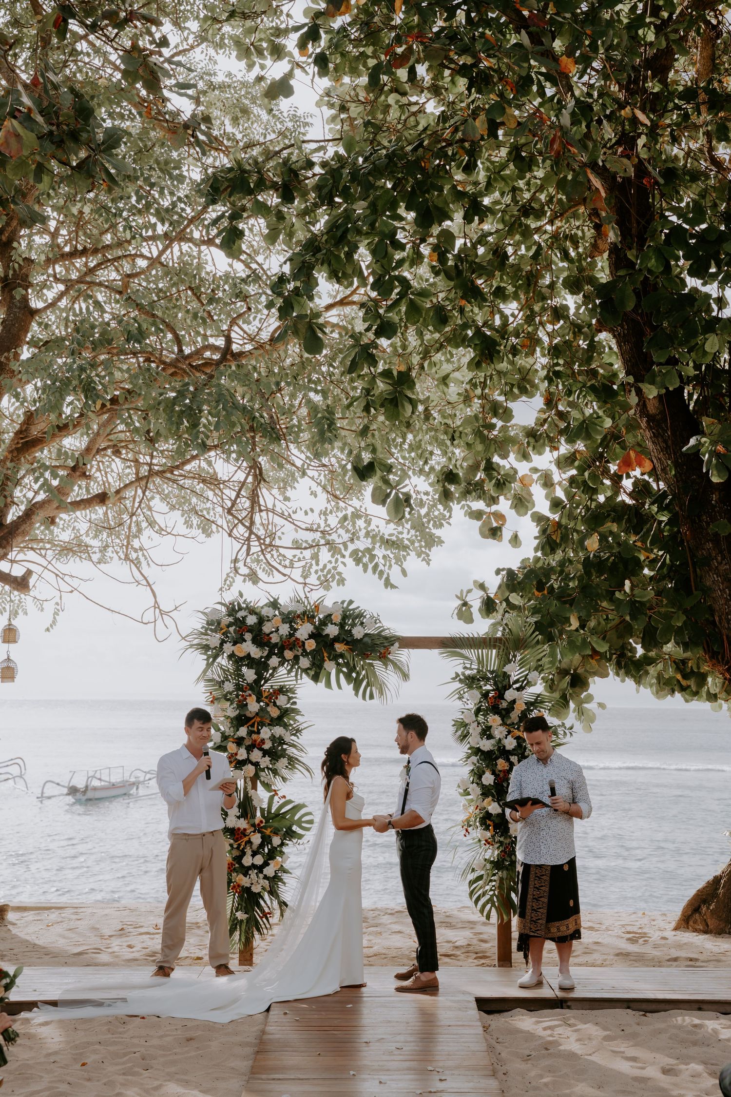A beachside wedding ceremony takes place under a tree canopy with the ocean in the background.
