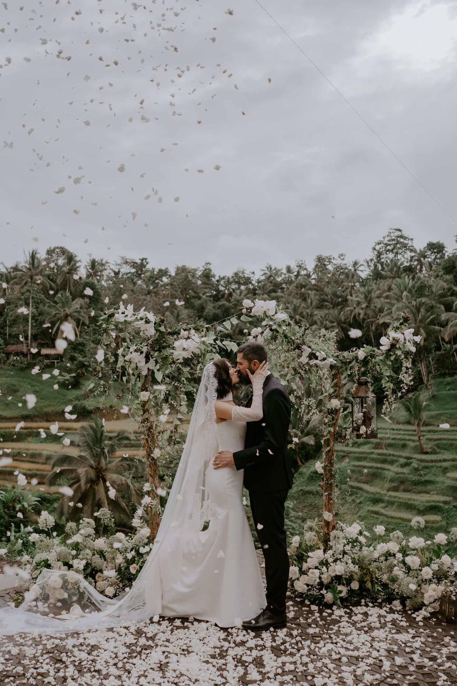 A couple in wedding attire stands in an orchard of blooming white cherry trees on their special day.