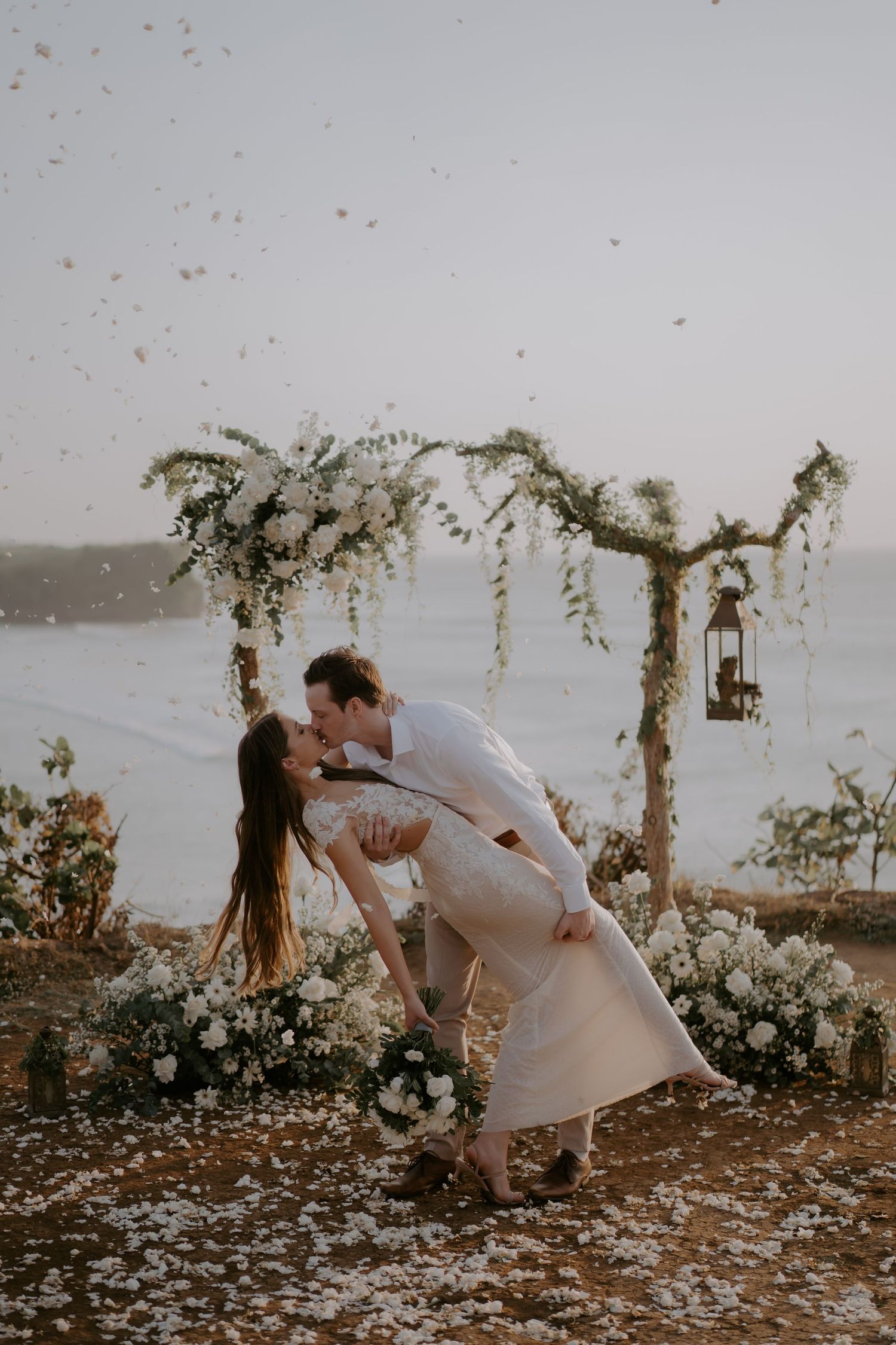 A romantic couple dances and dips during their wedding ceremony on a flower-covered beach at sunset.