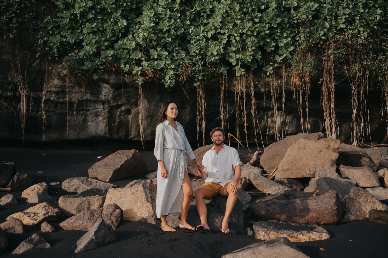 A couple sits together on large rocks near a wooded area during golden hour sunlight.