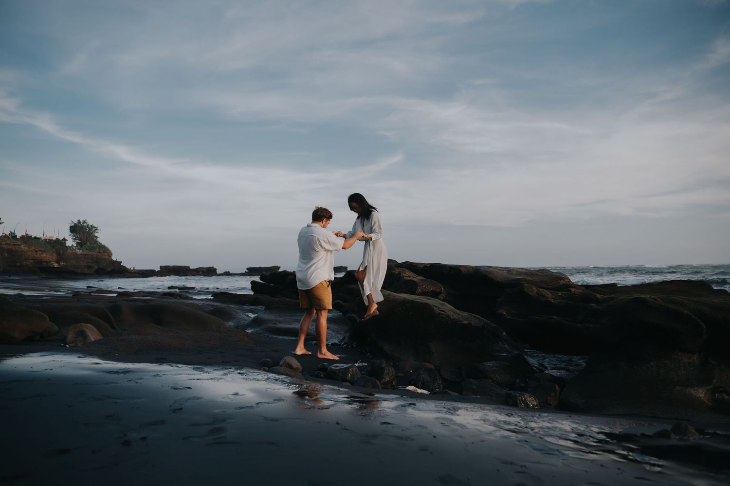 A couple shares a romantic moment on rocky coastal cliffs overlooking the ocean at sunset.