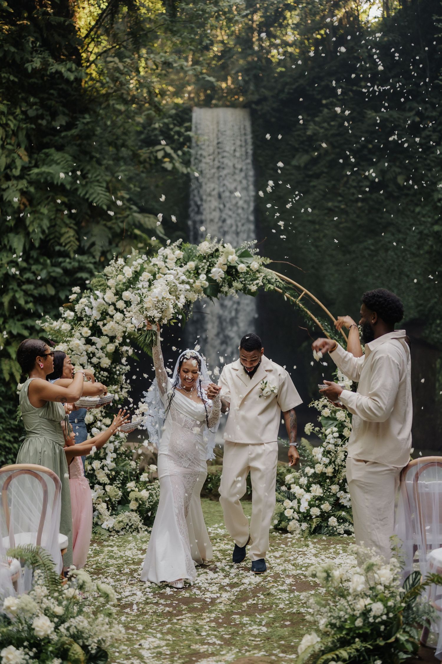 Joyful newlyweds walk down the aisle as guests toss white flower petals in a tropical forest setting.