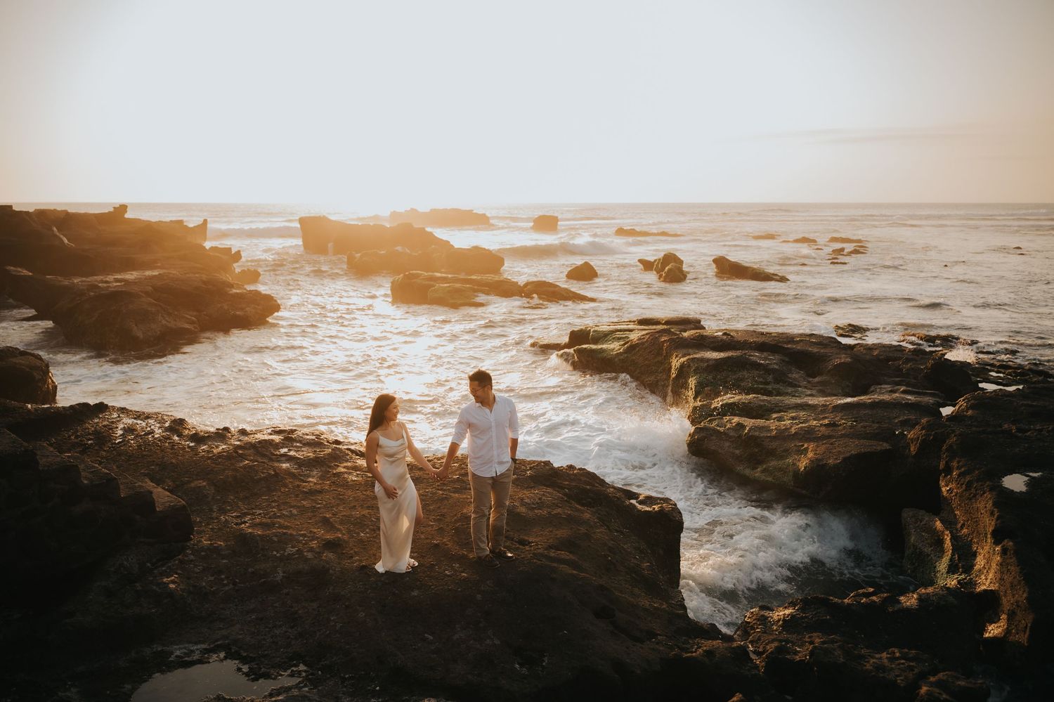 Couple holds hands on rocky coastal cliffs at golden sunset with ocean waves crashing around them.