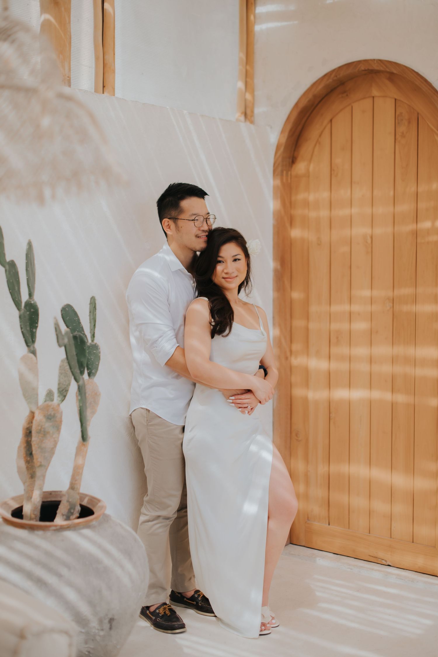 A couple poses together in a bright boho studio with a wooden arched door and cactus, woman in white dress.