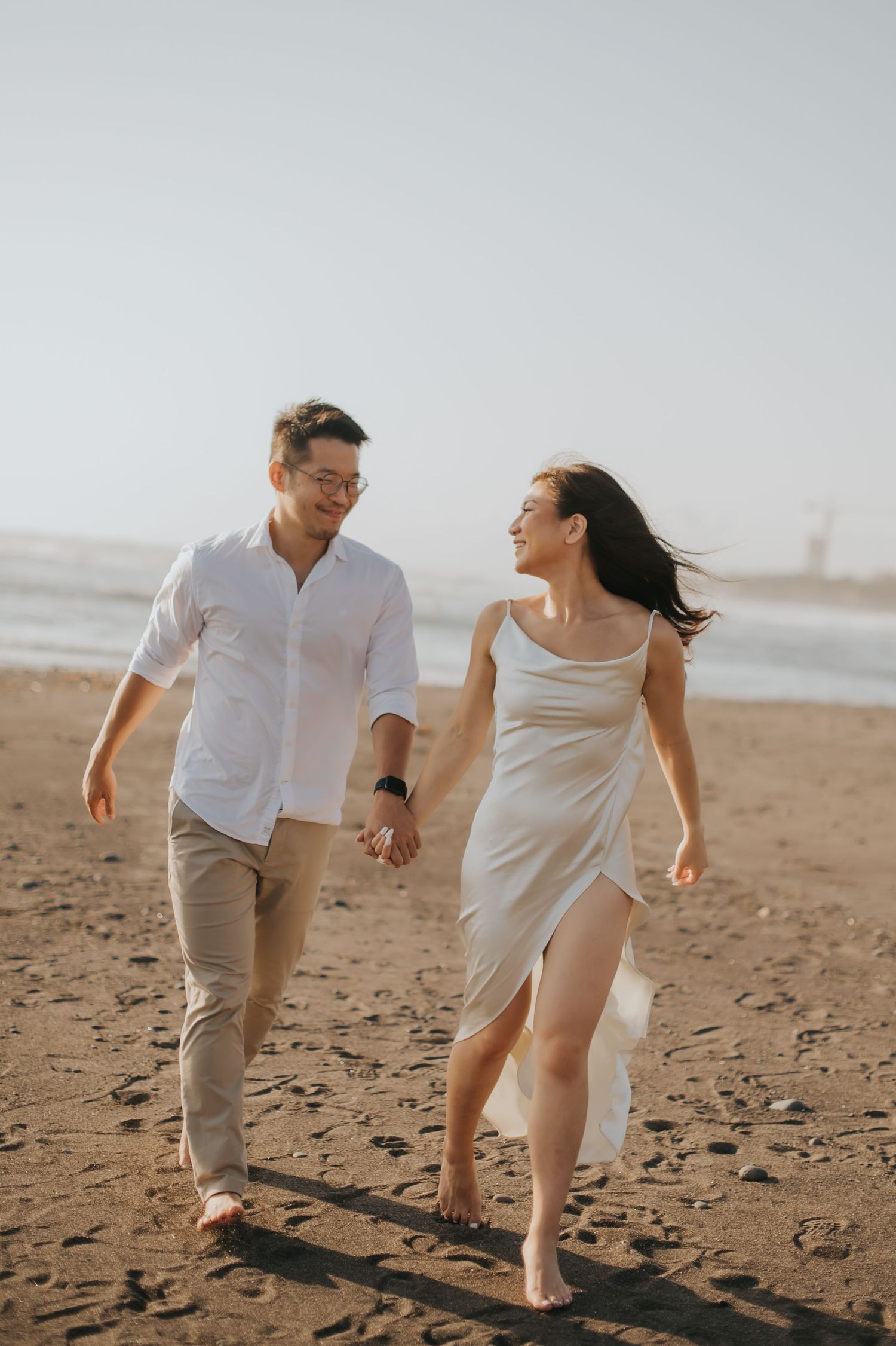 Couple holding hands and walking barefoot on a sandy beach, smiling at each other in white outfits.