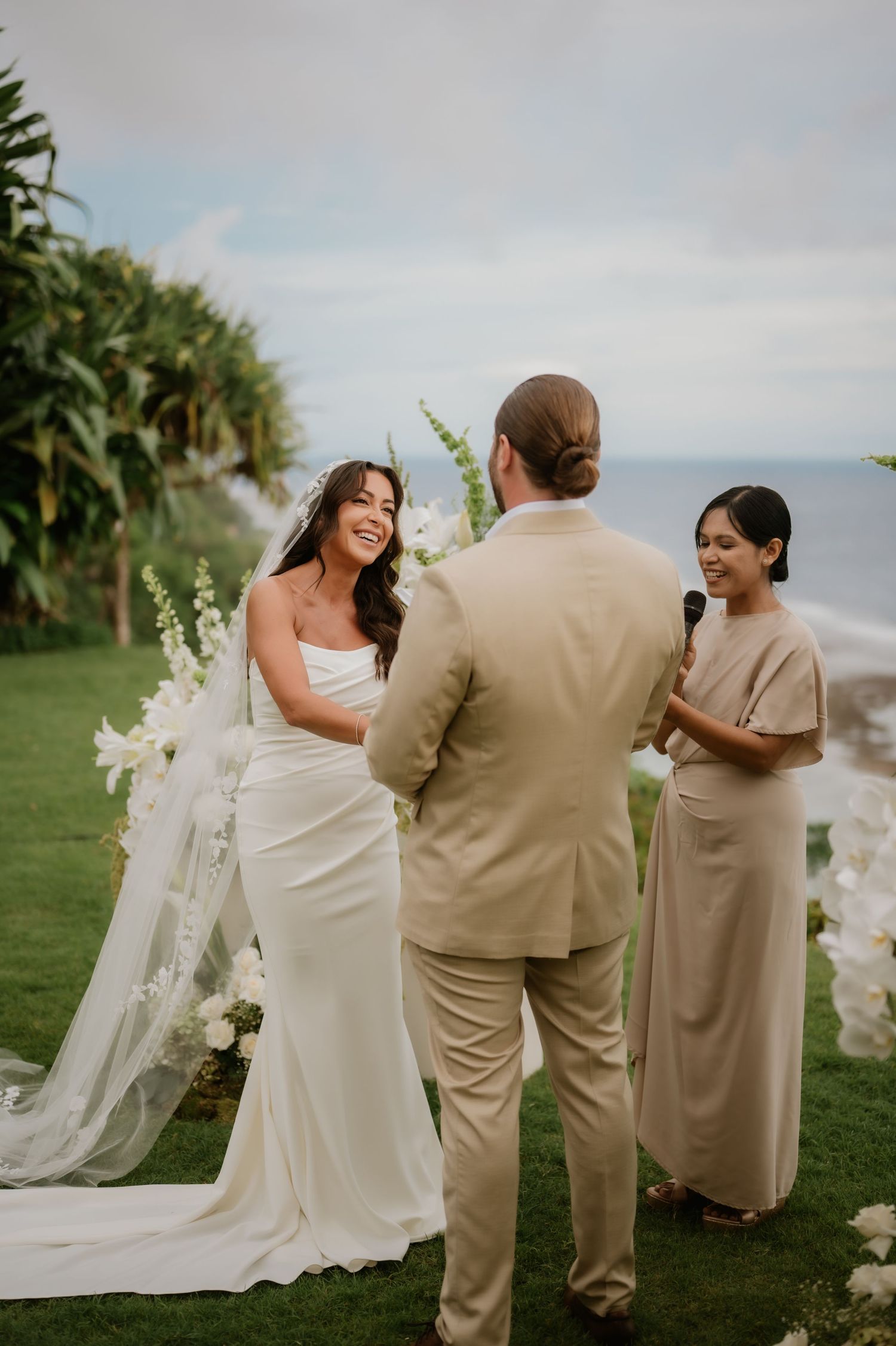 Bride in white gown laughs joyfully during outdoor wedding ceremony as officiant speaks.