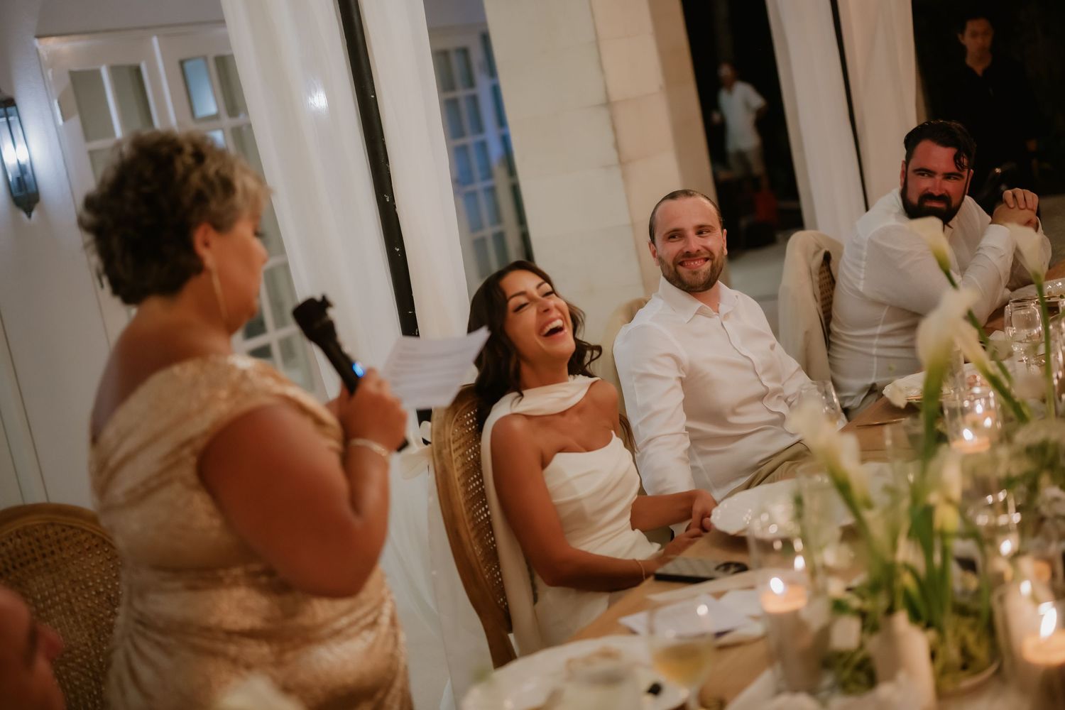 Bride and groom laughing joyfully during heartfelt wedding speech at candlelit reception dinner table.