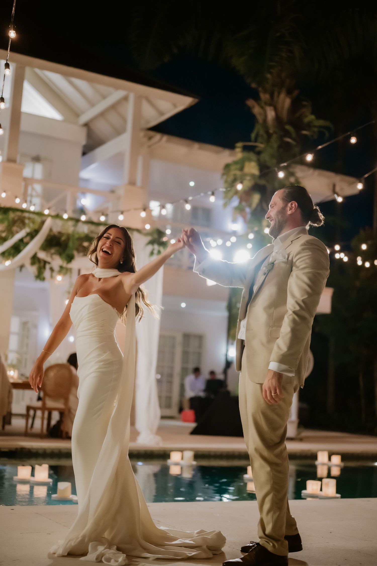 Laughing bride in white dress dancing with groom in tan suit beside candle-lit pool at night.