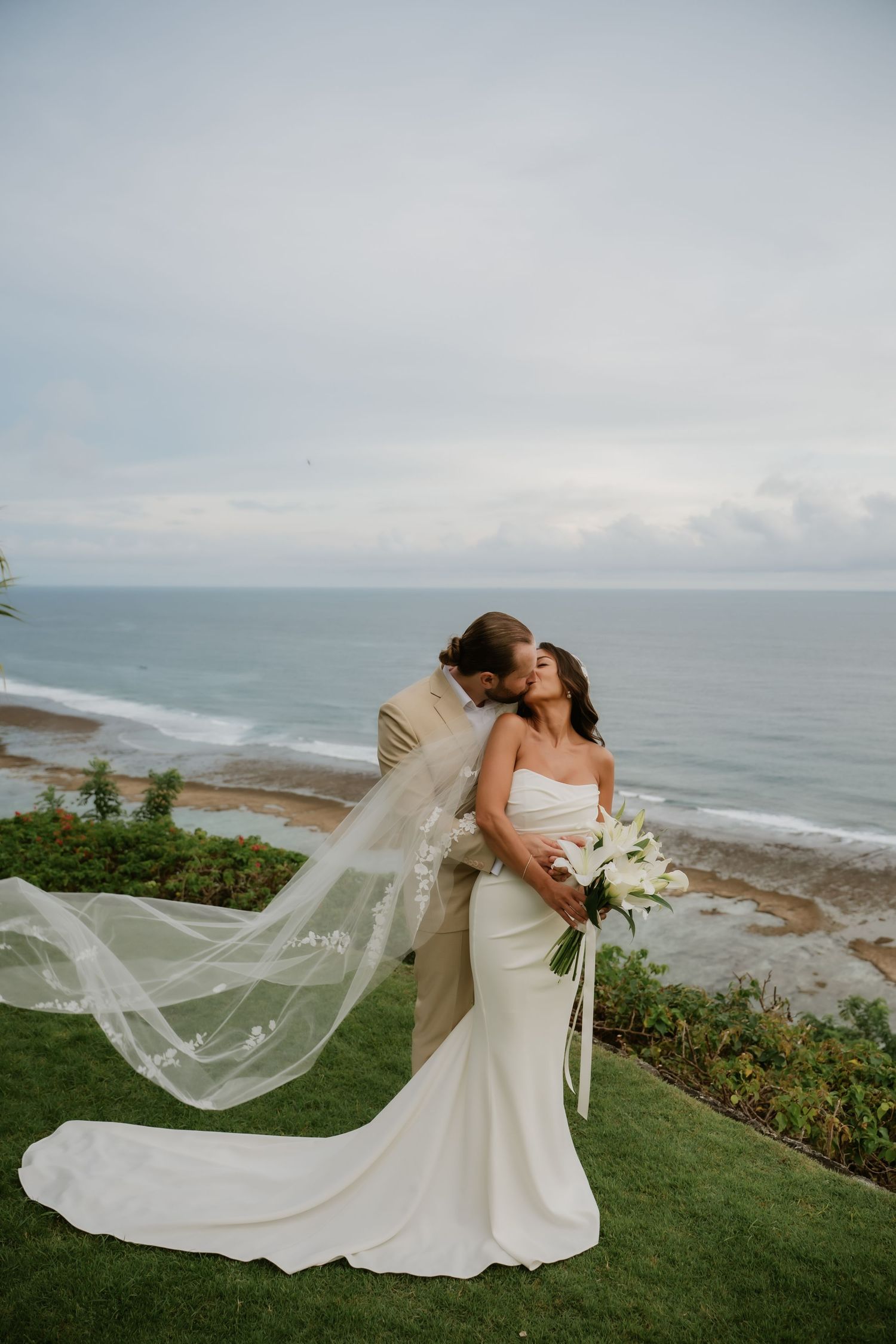 Bride and groom share a romantic kiss overlooking the ocean, her veil flowing in the wind at a coastal wedding.