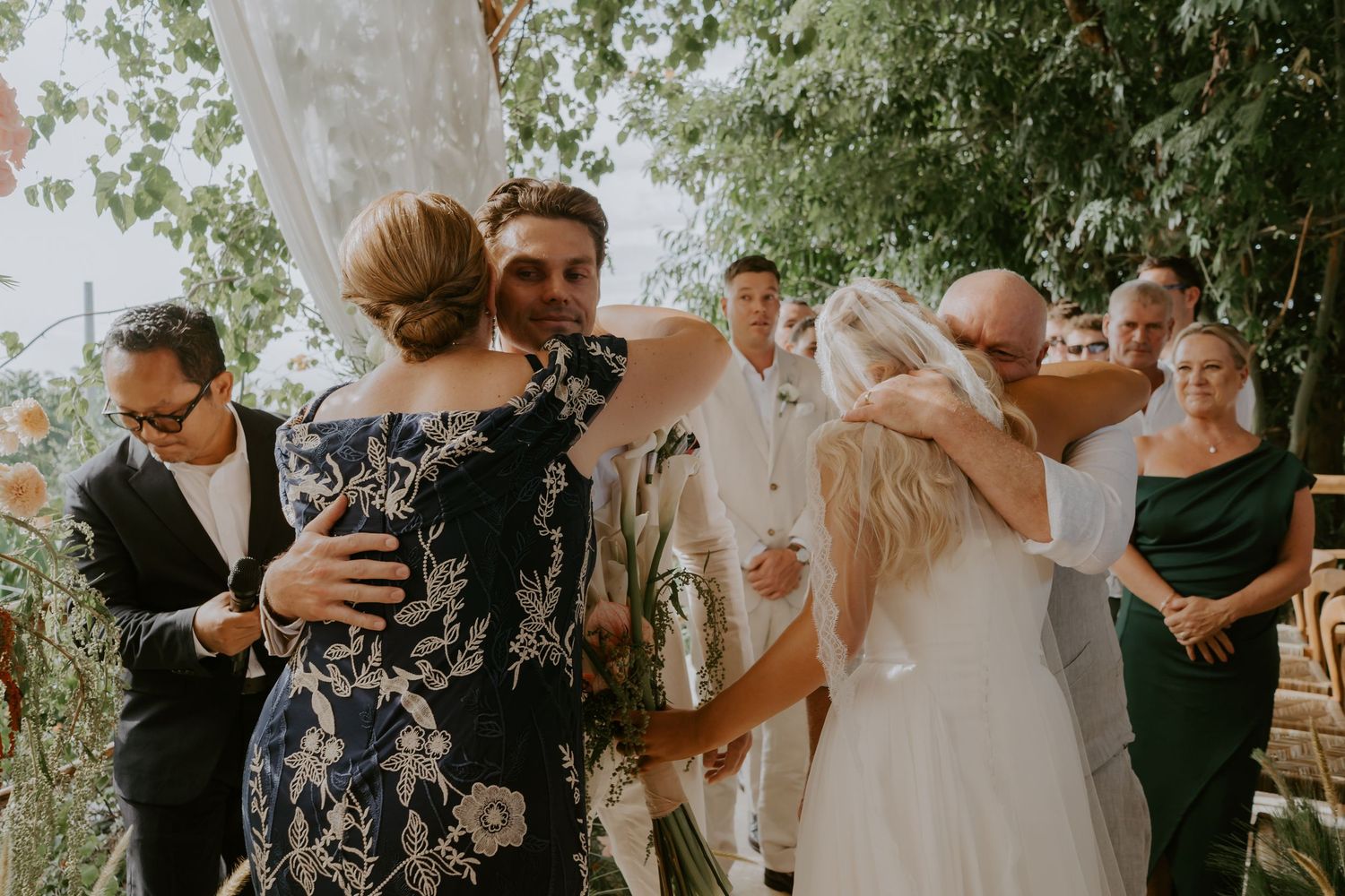 Joyful wedding ceremony moment with bride and groom sharing hugs with family members outdoors.