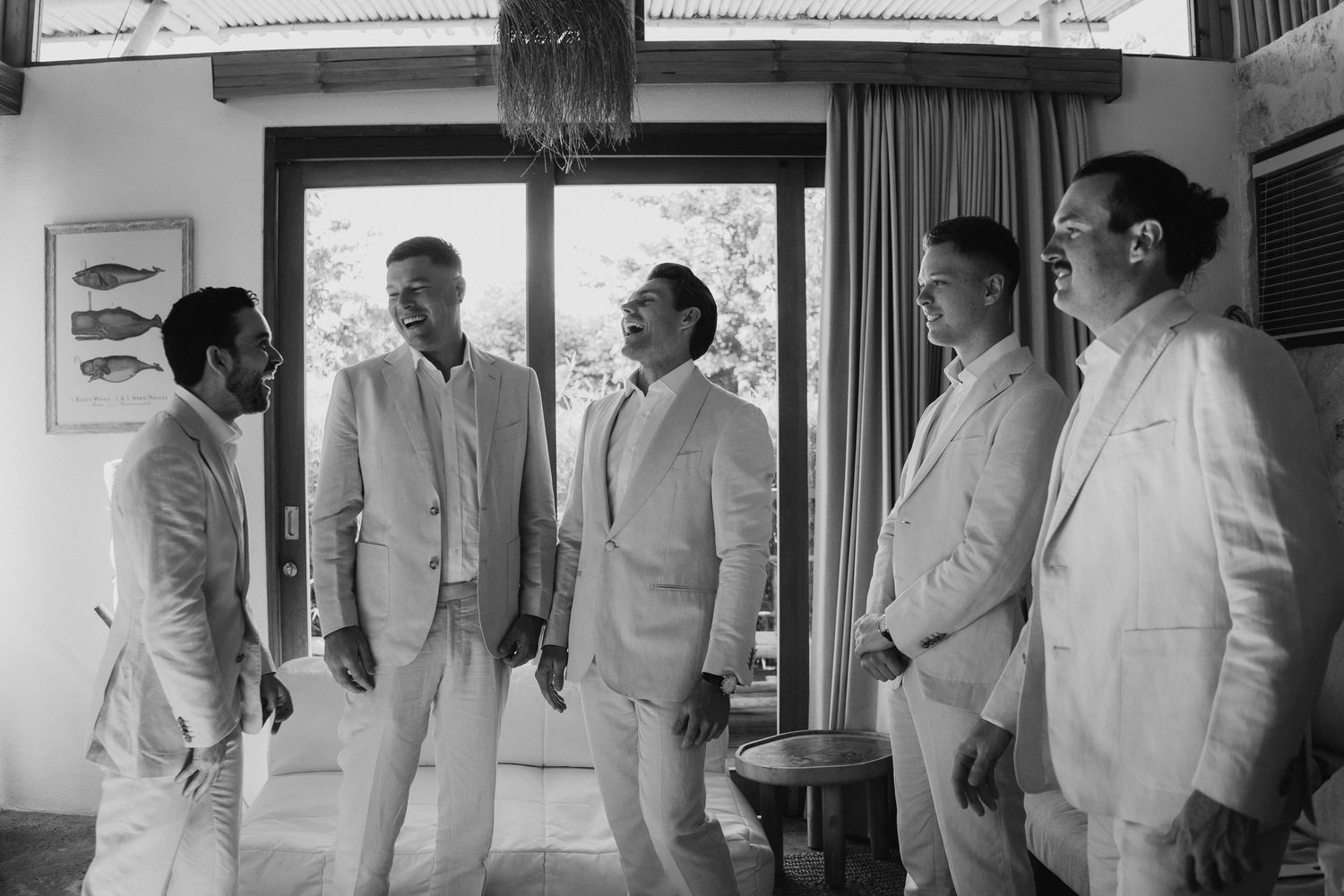 Five groomsmen in matching white linen suits laughing together in a tropical villa in a black and white photo.