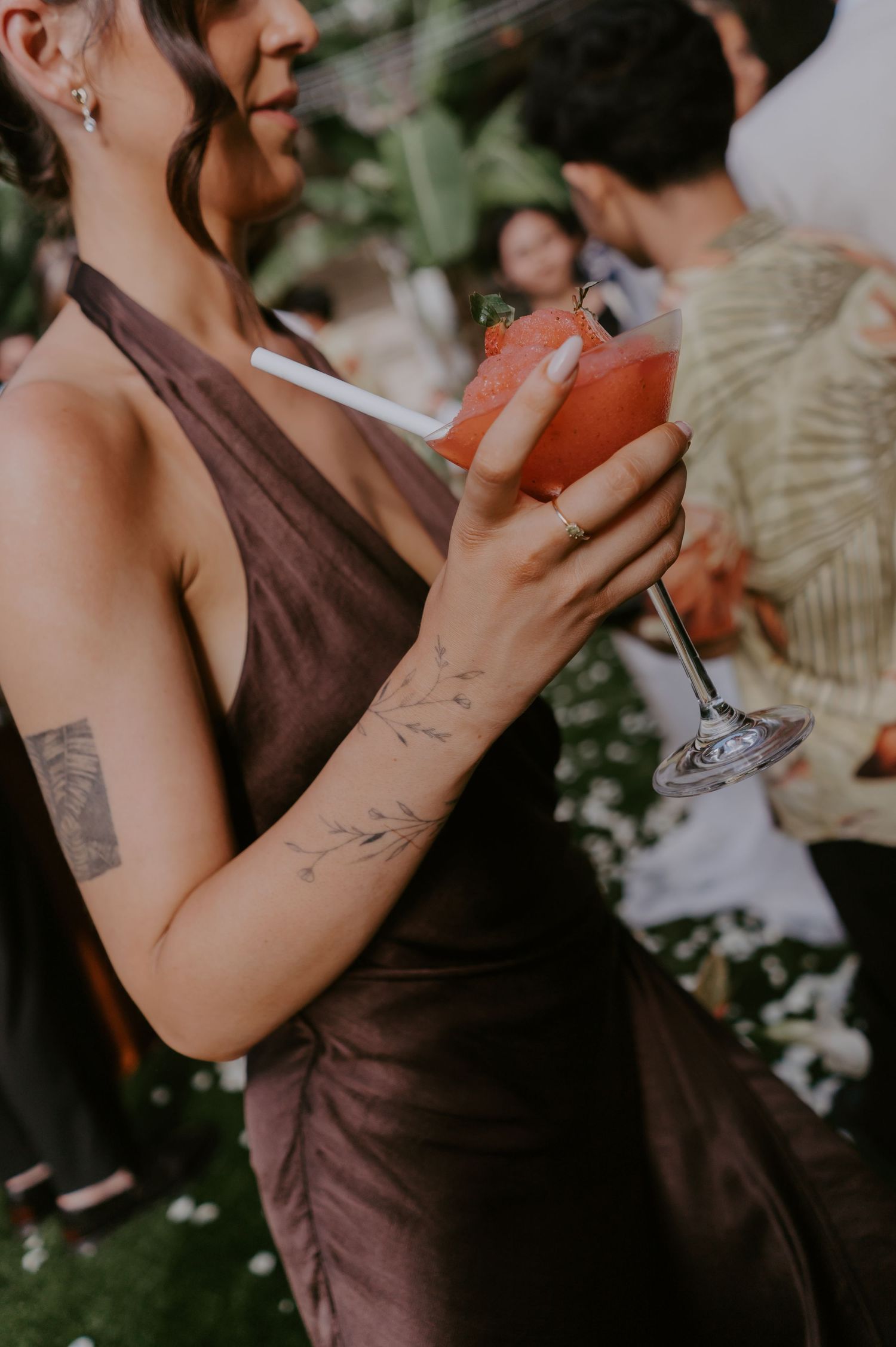 Wedding guest in brown halter dress holds a pink cocktail, showcasing delicate floral arm tattoo.