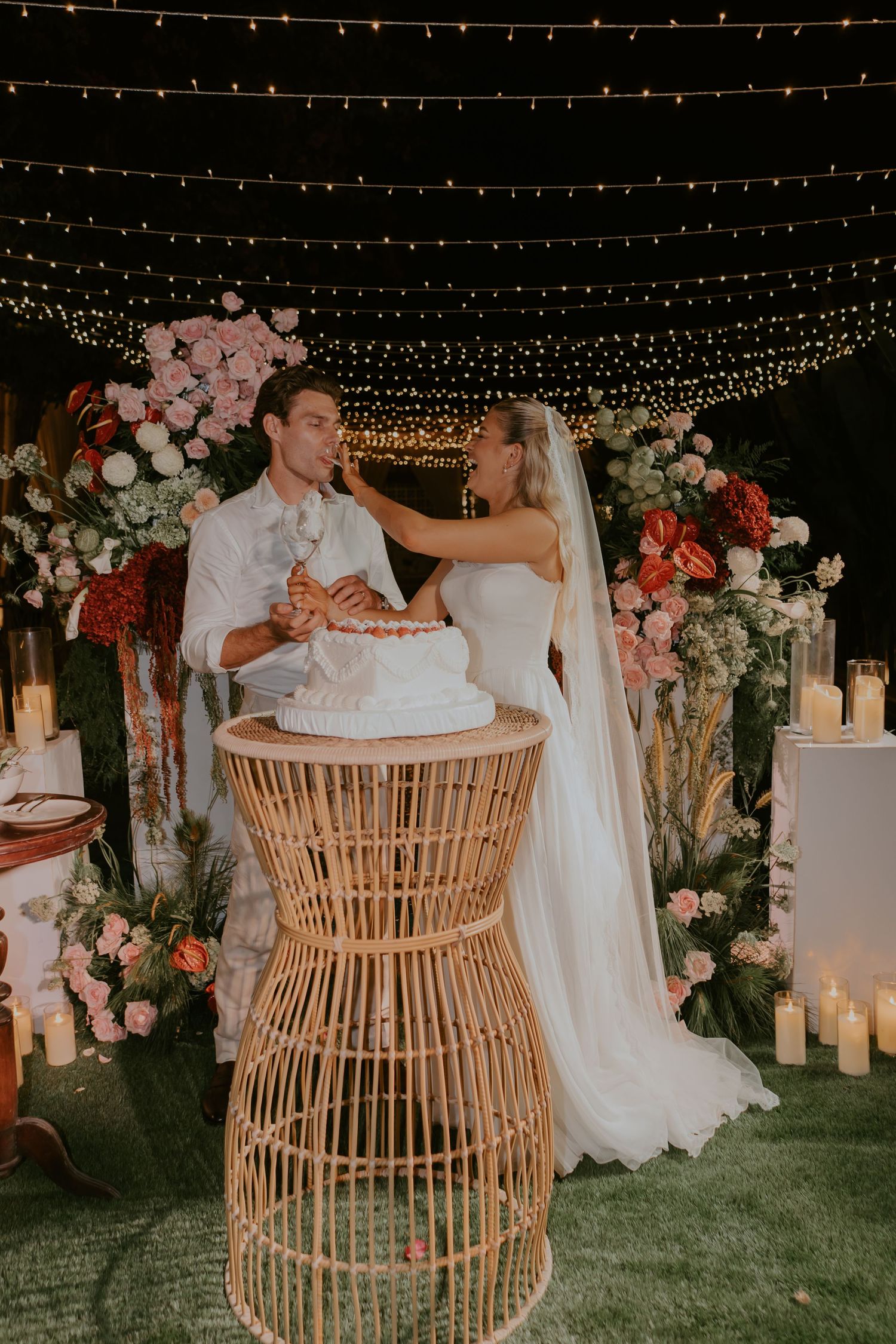 Bride and groom cutting their white wedding cake on a rattan stand surrounded by candles and floral arrangements at night.