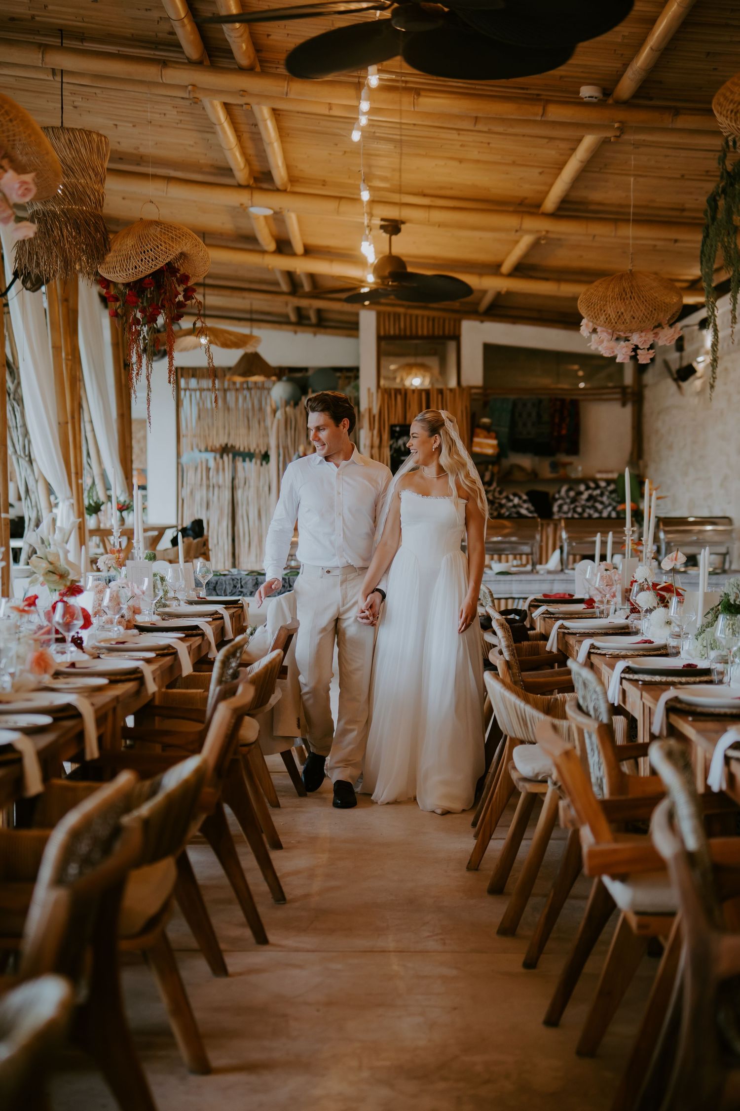 Bride and groom walk hand in hand through elegantly decorated bamboo wedding reception hall.